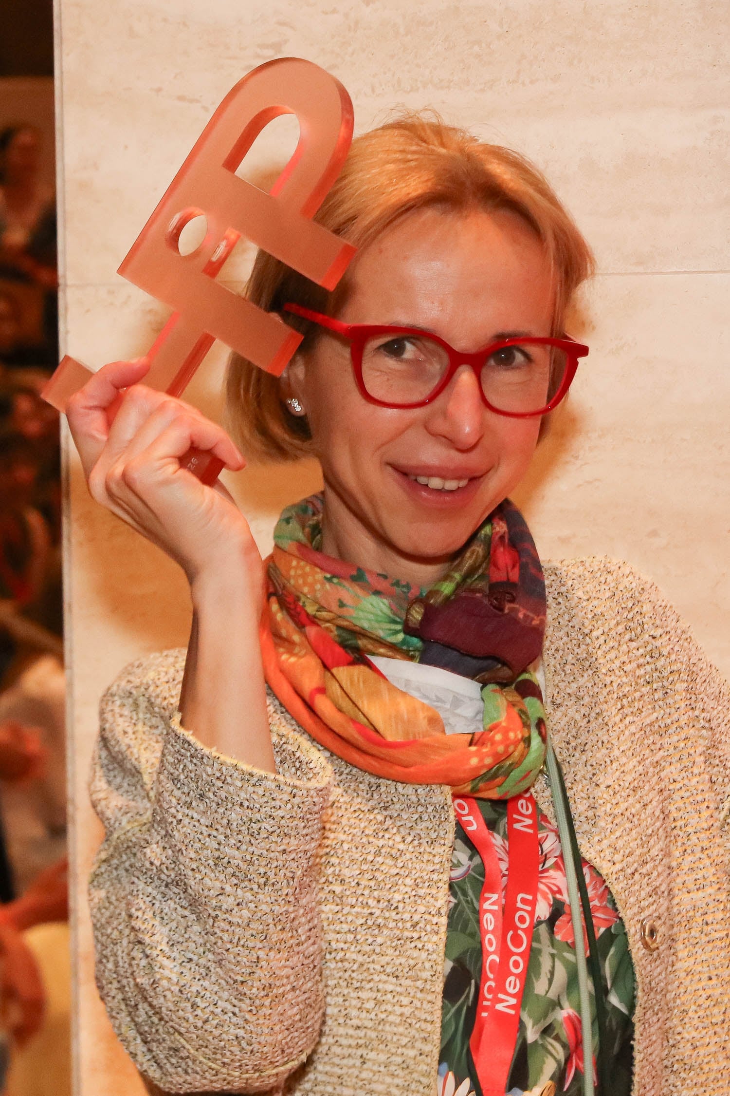 A woman wearing red glasses and a colorful scarf holds a translucent red award shaped like the letter "P" beside her head.