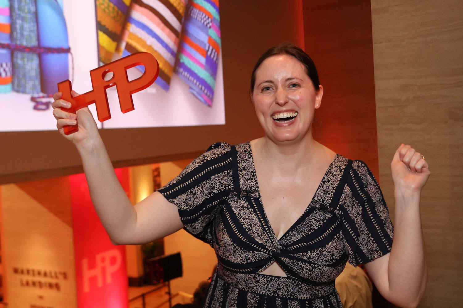 A woman smiles and raises an orange "HiP" sign at an indoor event, with a colorful textile image projected on the wall behind her.