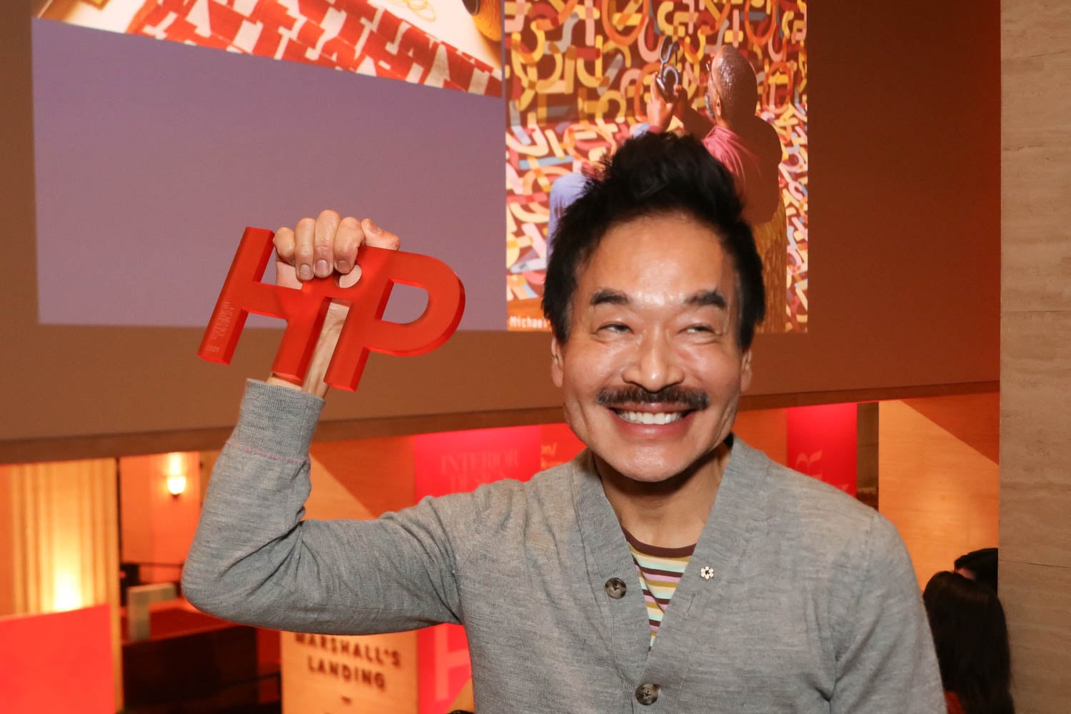 A person smiling and holding up a red "HIP" sign, with colorful artwork and event displays in the background.