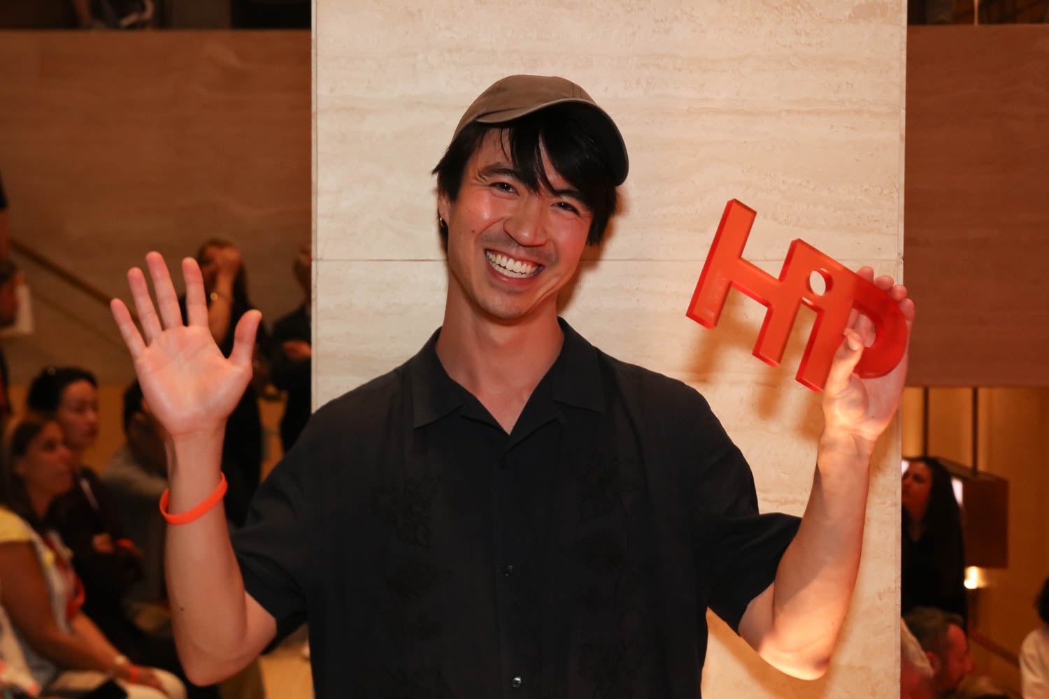 A person wearing a black shirt and cap smiles at the camera, holding up a red "HiD" sign and waving, with people seated in the background.