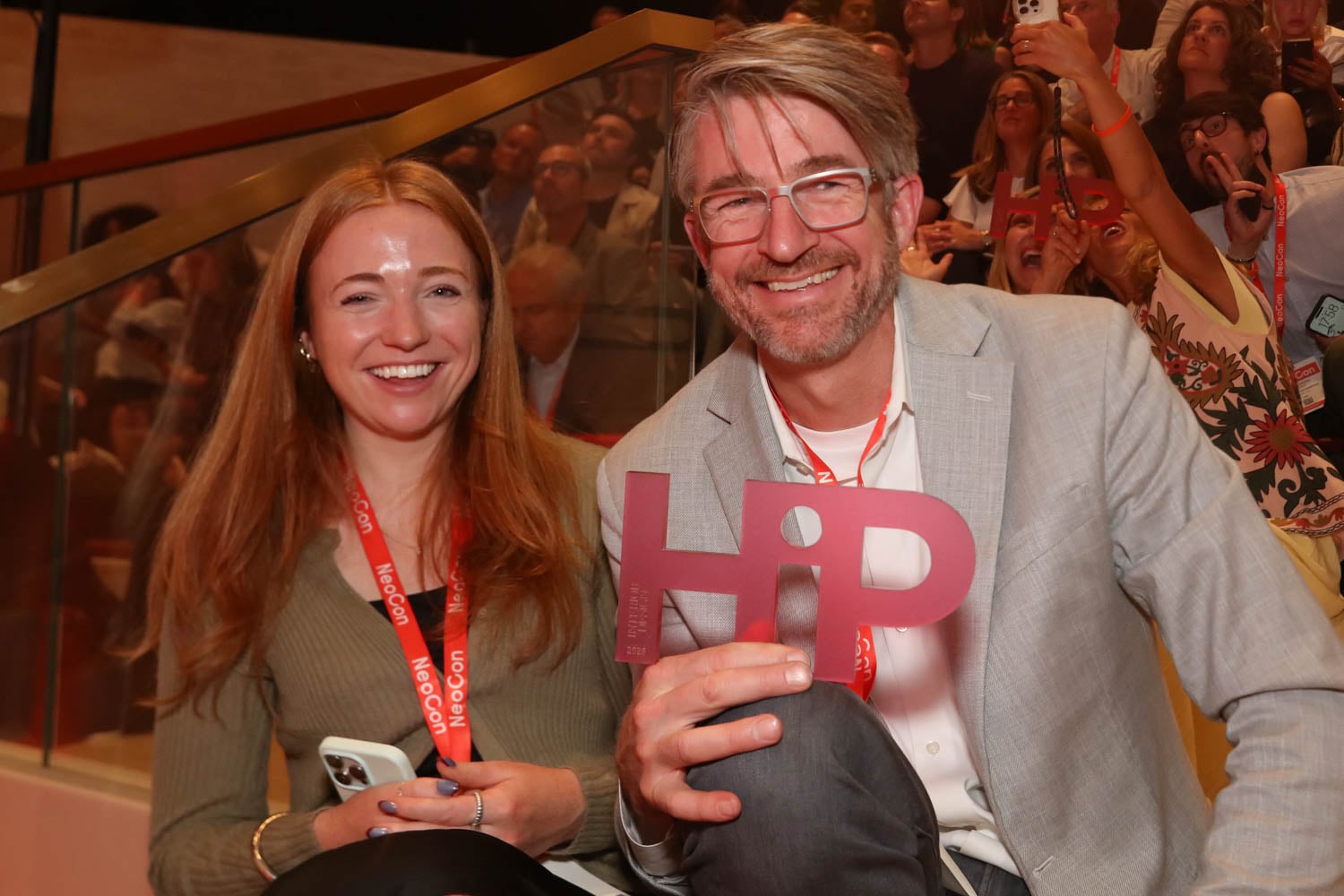 Two people sit smiling at an event, wearing red lanyards. The man holds a large "HIP" sign. A crowd is visible in the background.