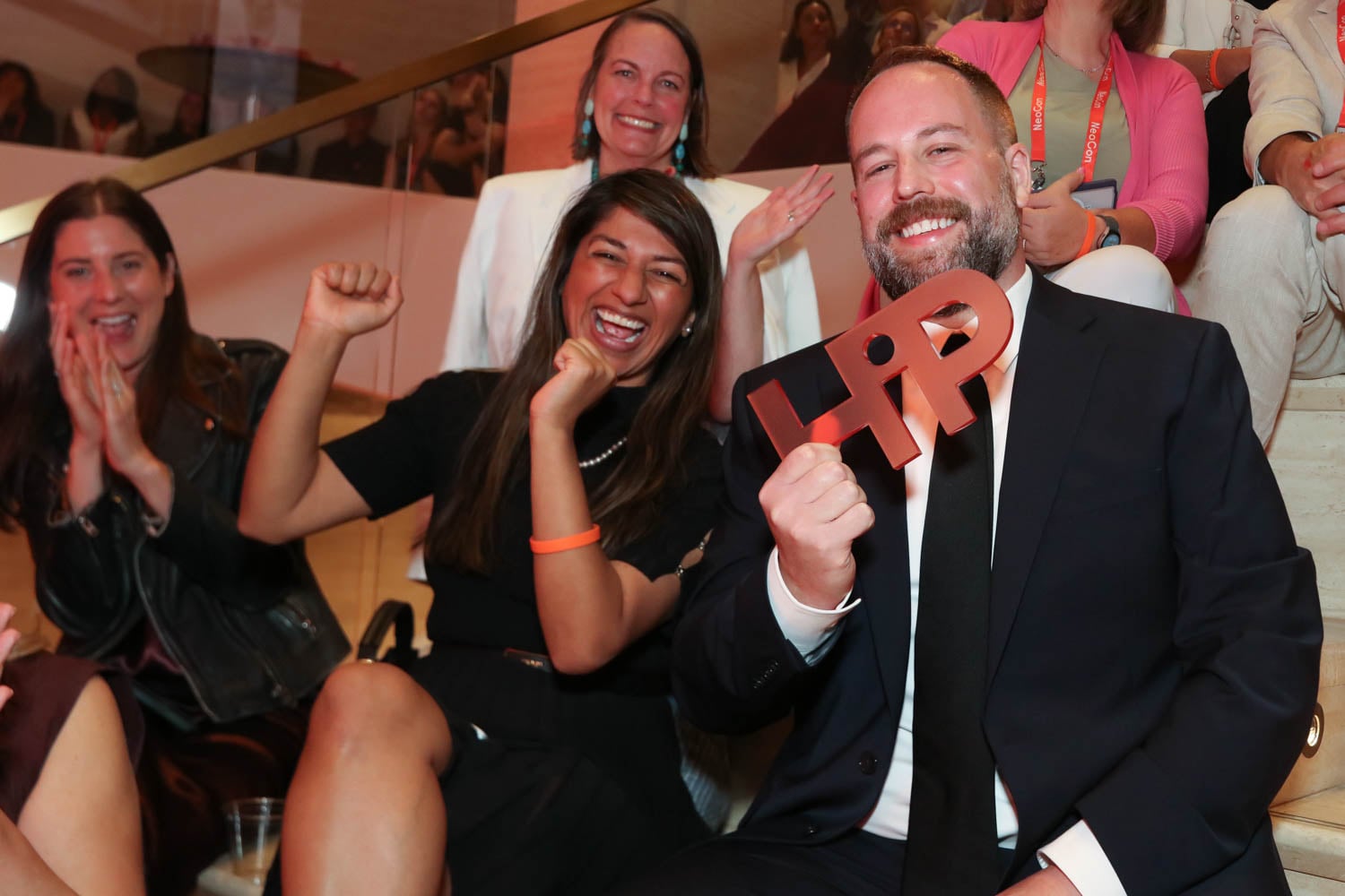 A group of people sitting on stairs, smiling and cheering; one man in a suit holds a red "HPP" cutout.