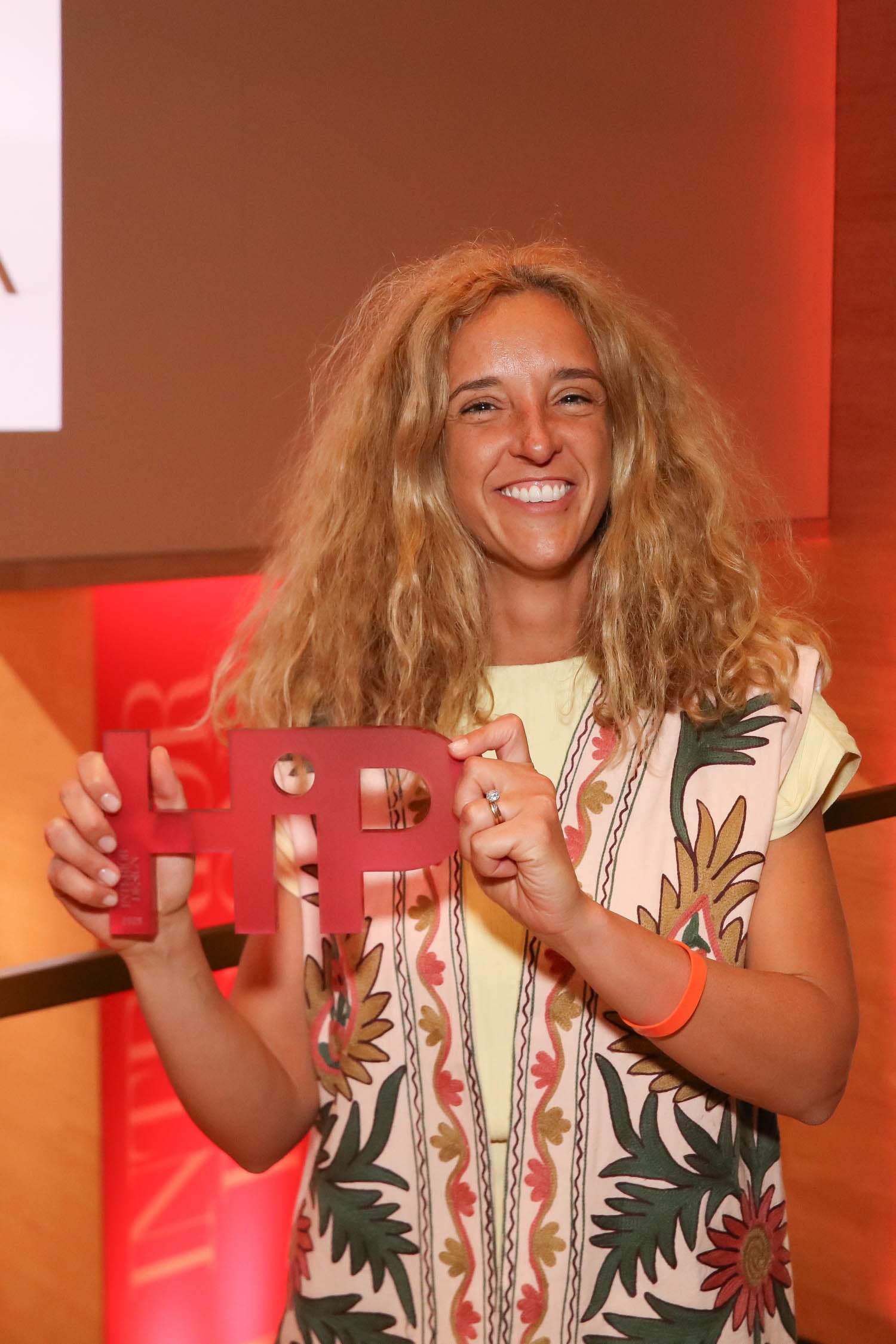 A person with curly blonde hair smiles while holding a red award shaped like the letters "HP" in front of a red-lit background.