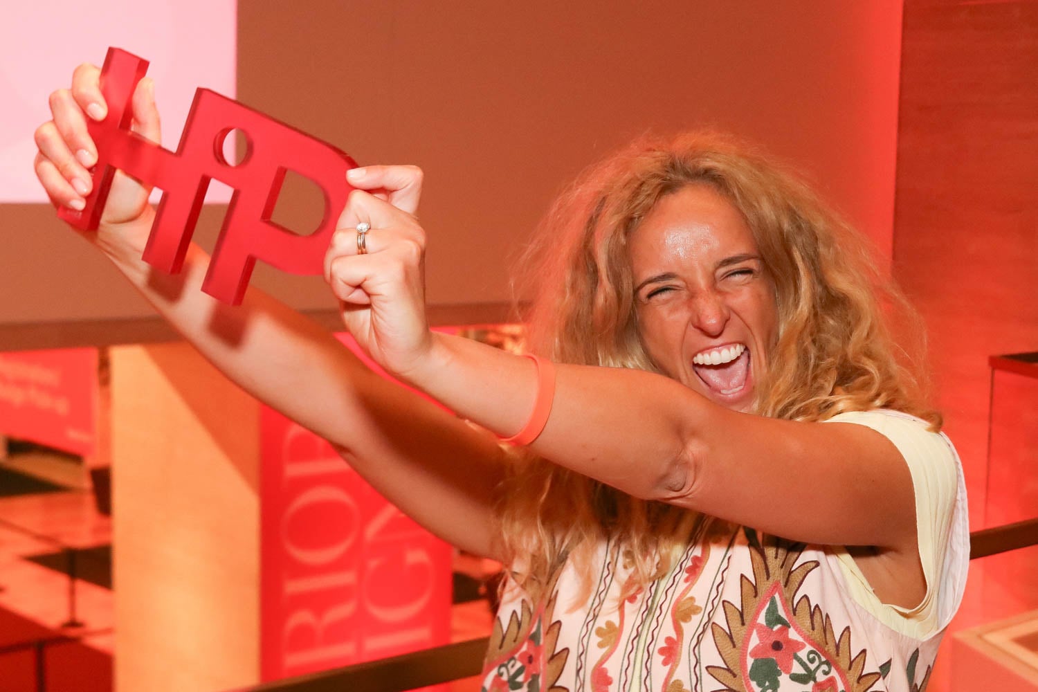 A woman with long blond hair smiles energetically while holding up a large red "HIP" sign in a brightly lit indoor space with red lighting.