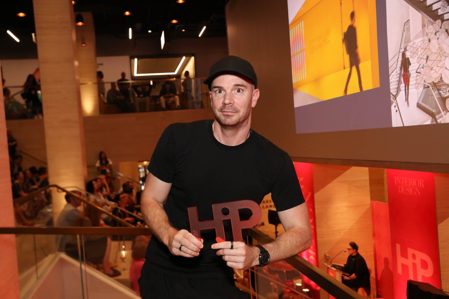 A man in a black t-shirt and cap holds an HP-shaped award in a modern, well-lit venue with people in the background and design projections on the wall.