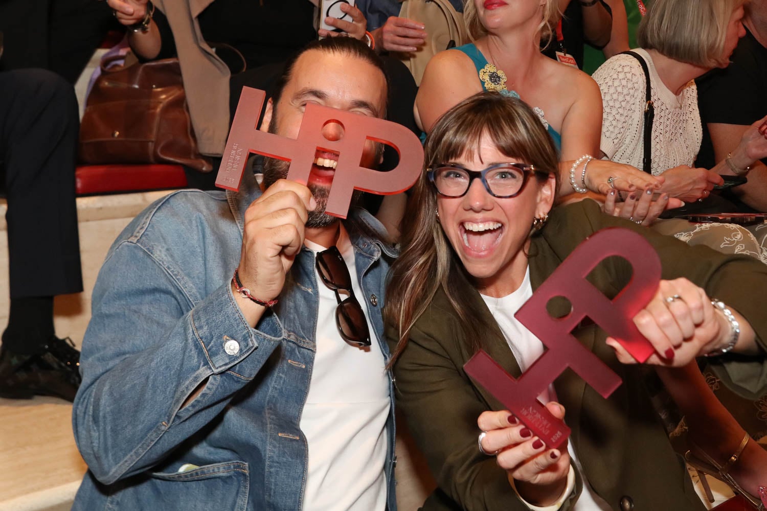 Two people sitting on stairs, smiling and holding up large red "HP" cutout signs at an indoor event with others in the background.