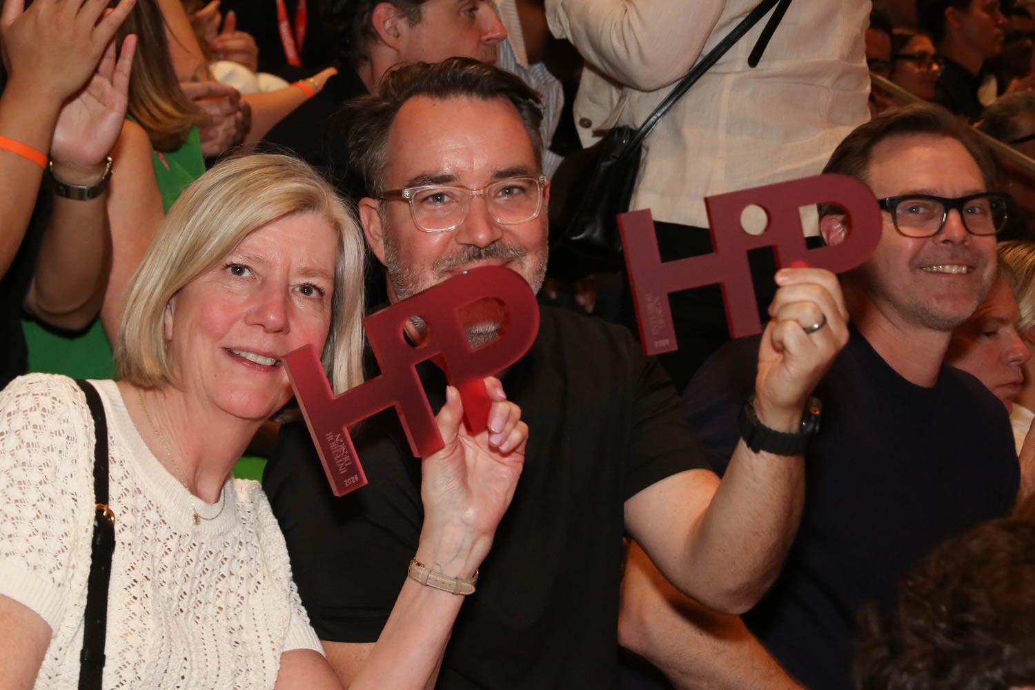 Three people in a crowd hold up large cut-out letters spelling "HiP" and smile toward the camera.