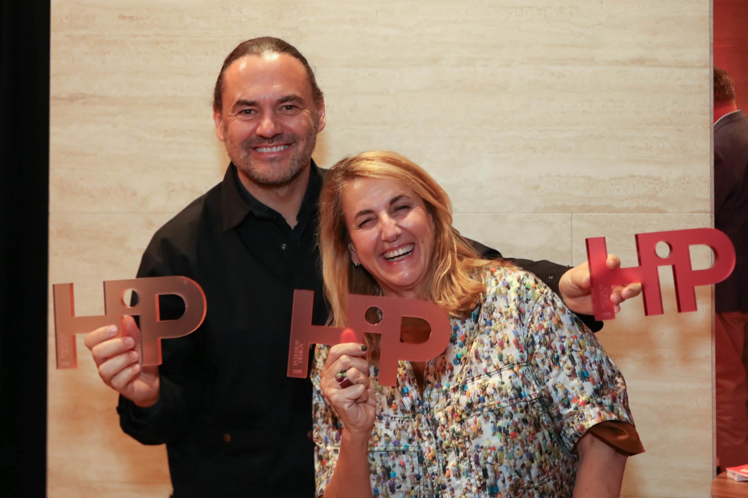 Two people smiling and holding large "HiP" award plaques while posing for a photo against a light-colored wall.