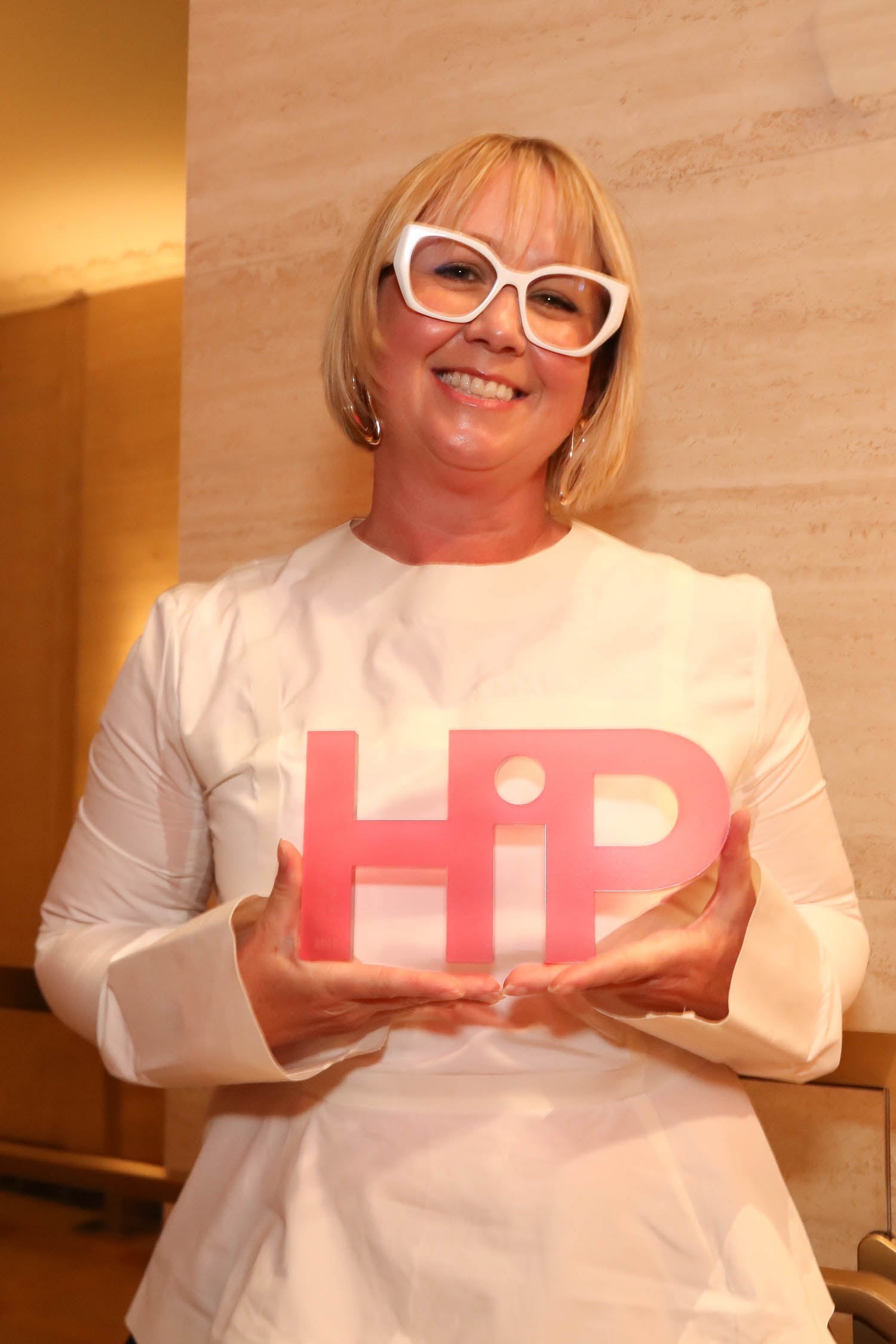 Smiling woman with short blonde hair and white glasses holds a pink award shaped like the word "HiP" in front of a beige wall.