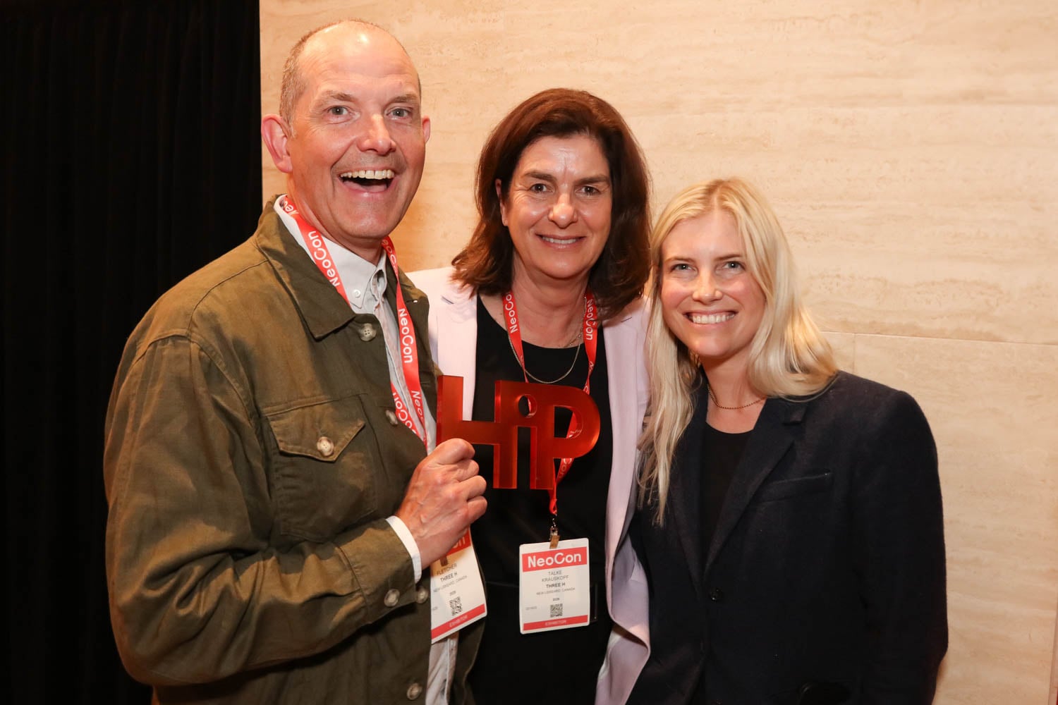 Three people wearing NeoCon badges pose together indoors; one holds a red "HiP" award and smiles at the camera.