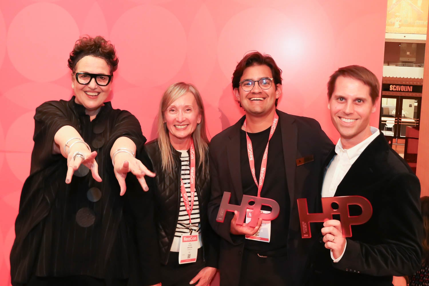 Four people stand smiling in front of a pink backdrop. Two are holding “HiP” awards. All wear event lanyards.