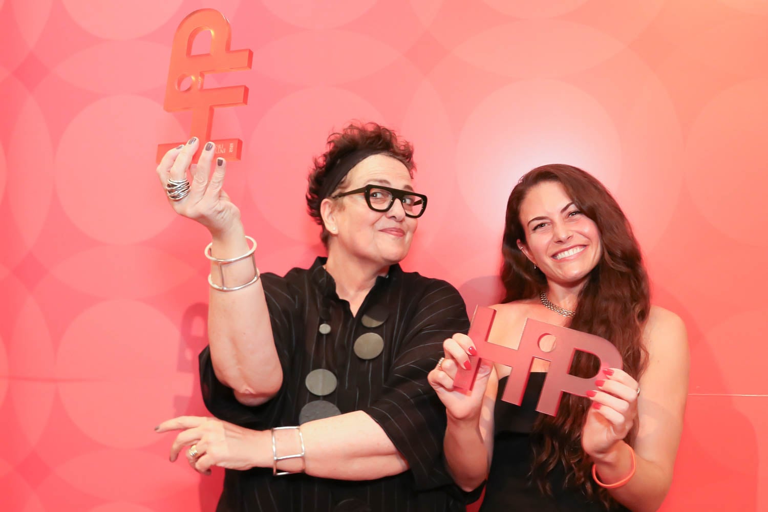 Two women stand in front of a red, patterned background, each holding large cutout letters spelling "HIP" and smiling at the camera.