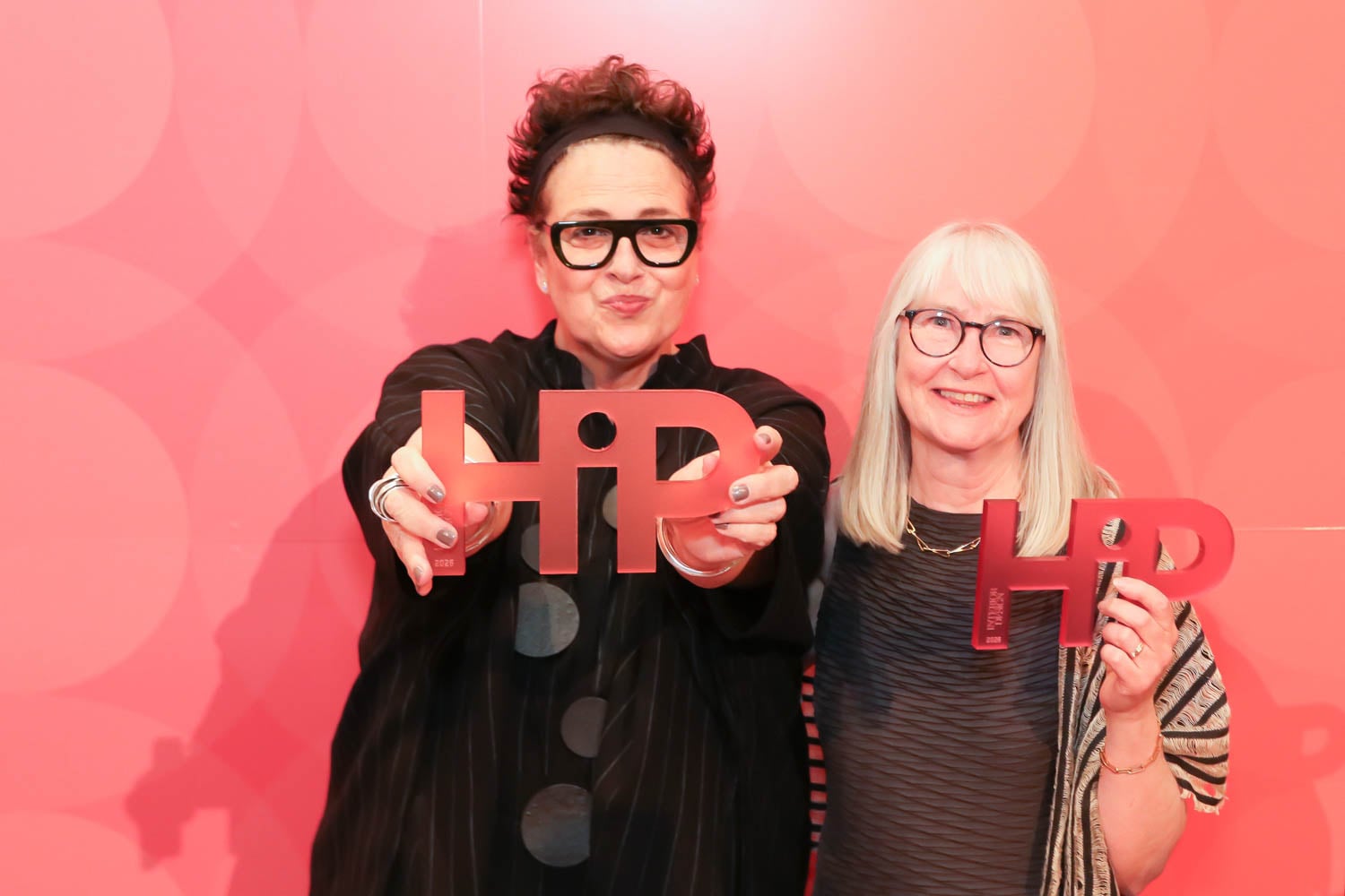 Two women stand in front of a red backdrop, smiling and holding metallic "HPD" signs toward the camera.