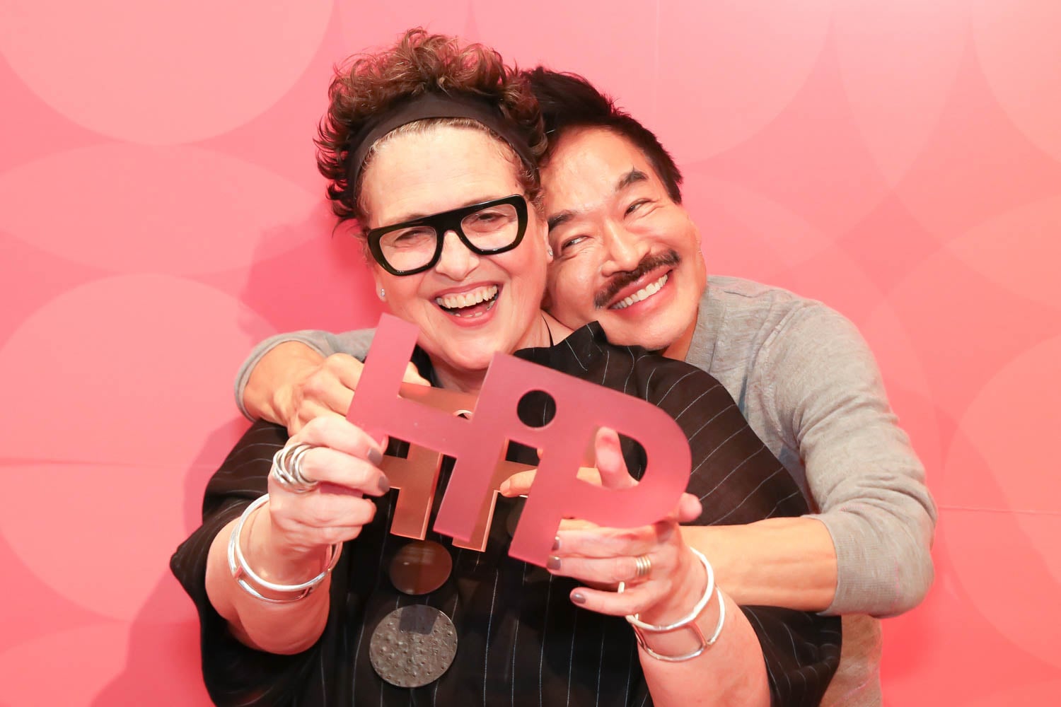 Two people smile and pose in front of a pink background, holding up a cutout sign that reads "HIP." The person in front wears glasses and multiple bangles.