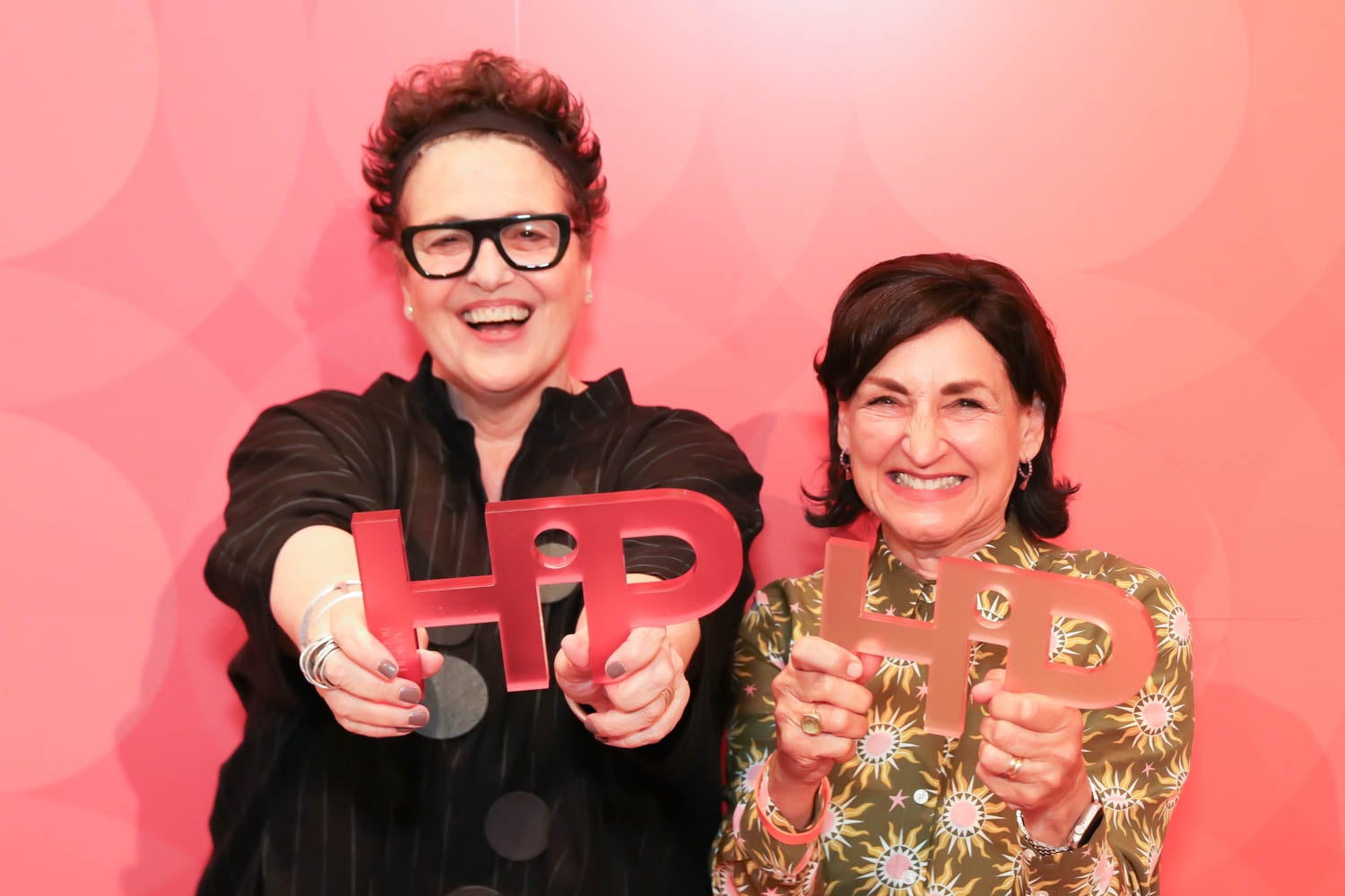 Two women smiling and holding HPD-shaped awards in front of a pink, patterned background.