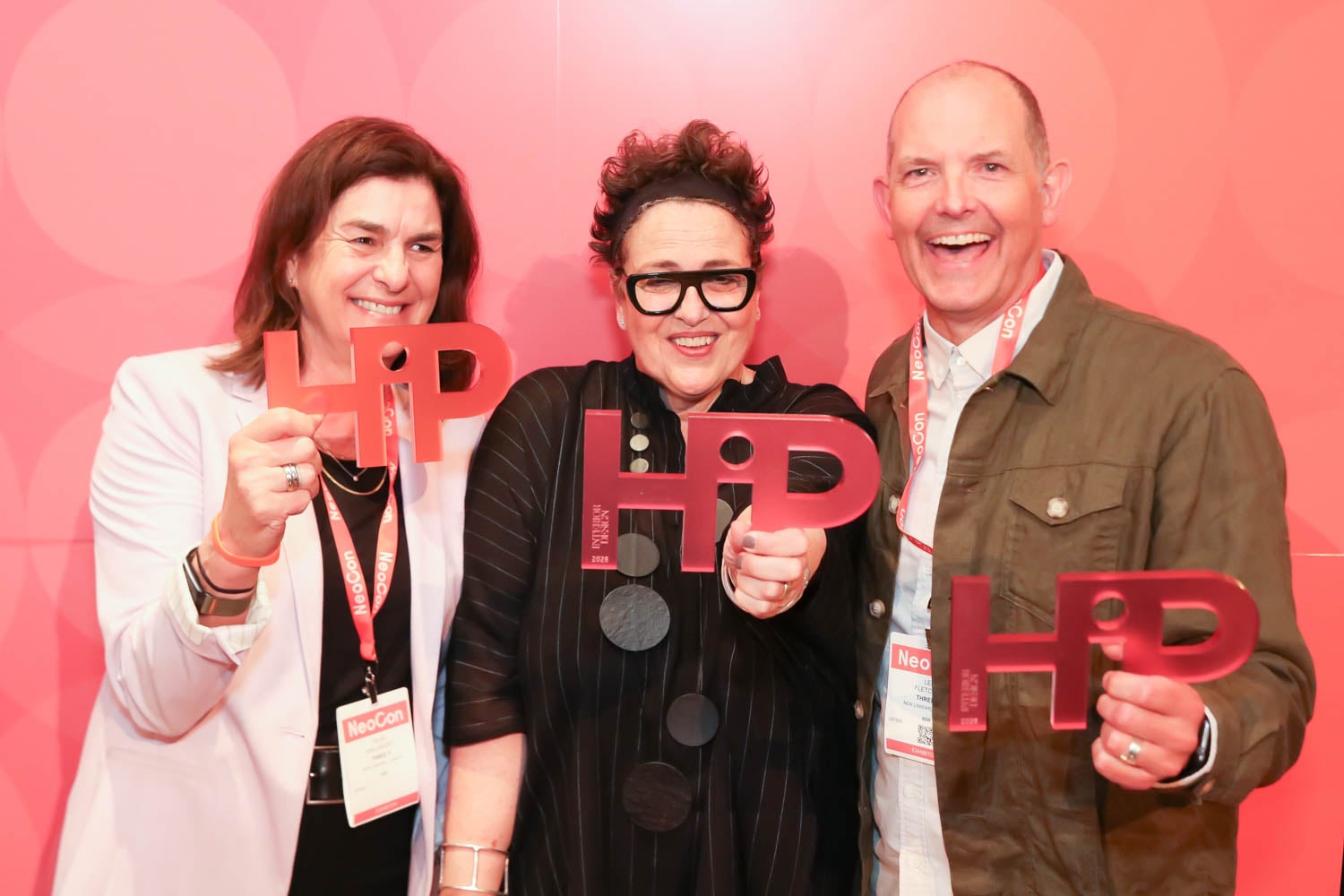 Three people smiling and holding red "HP" awards while standing in front of a red, circular-patterned backdrop.