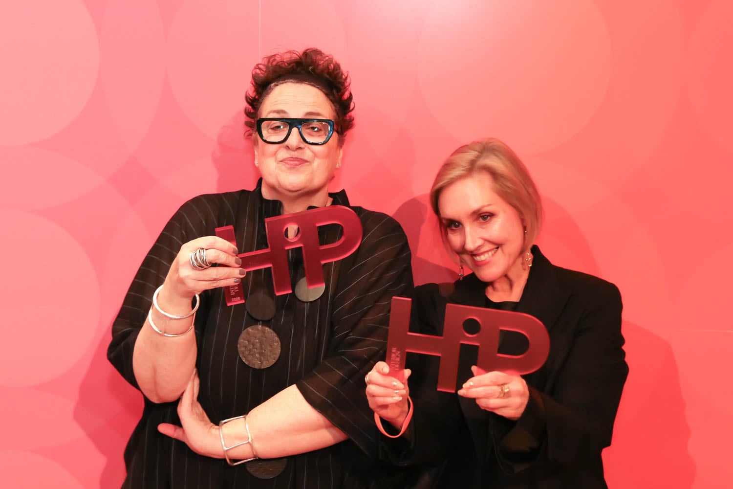 Two women stand in front of a pink backdrop, each holding a red "HIP" award and smiling at the camera.