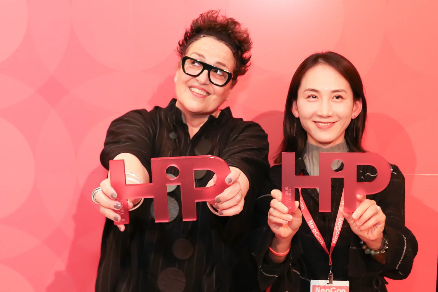 Two women stand in front of a red backdrop, smiling and holding "HiP" award trophies toward the camera.