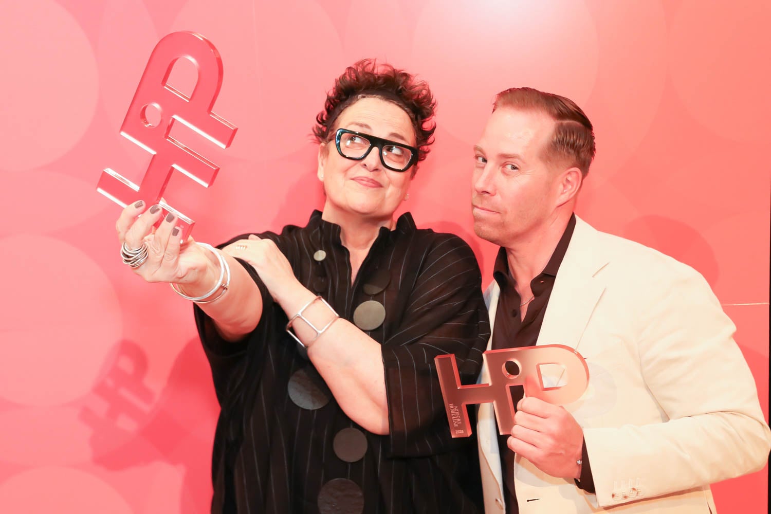 Two people stand in front of a red background, each holding a transparent award with "HIP" written on it. The person on the left is wearing glasses and a black outfit; the person on the right wears a light suit.