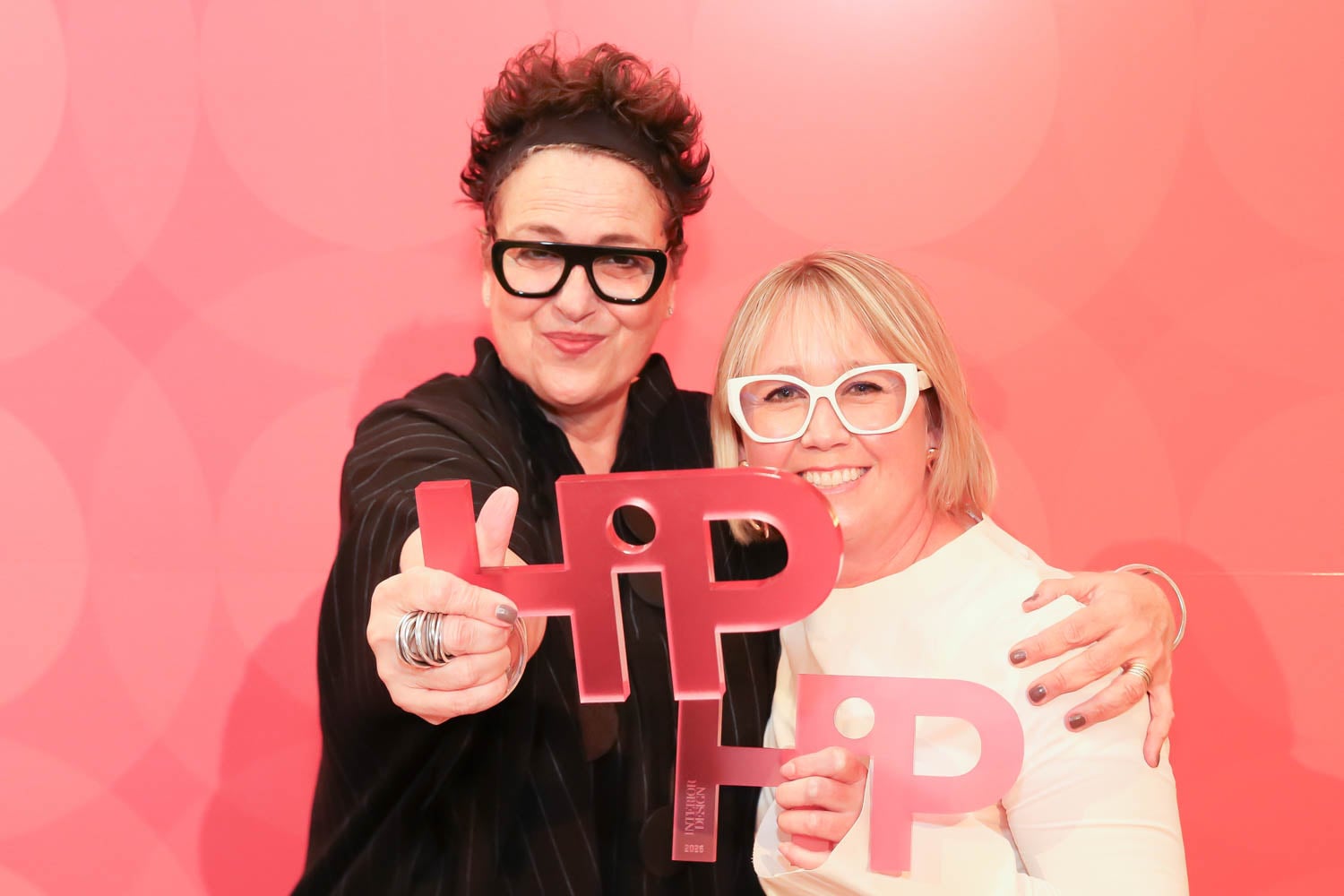 Two people wearing glasses smile and hold red "HIP" awards in front of a pink, patterned background.