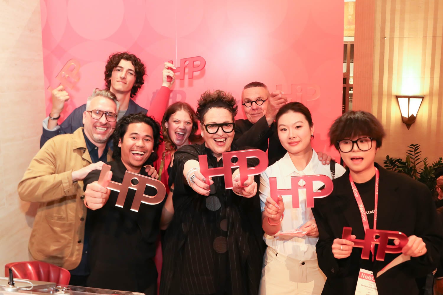 A group of eight people stand together indoors, smiling and holding red "HIP" signs in front of a pink backdrop.