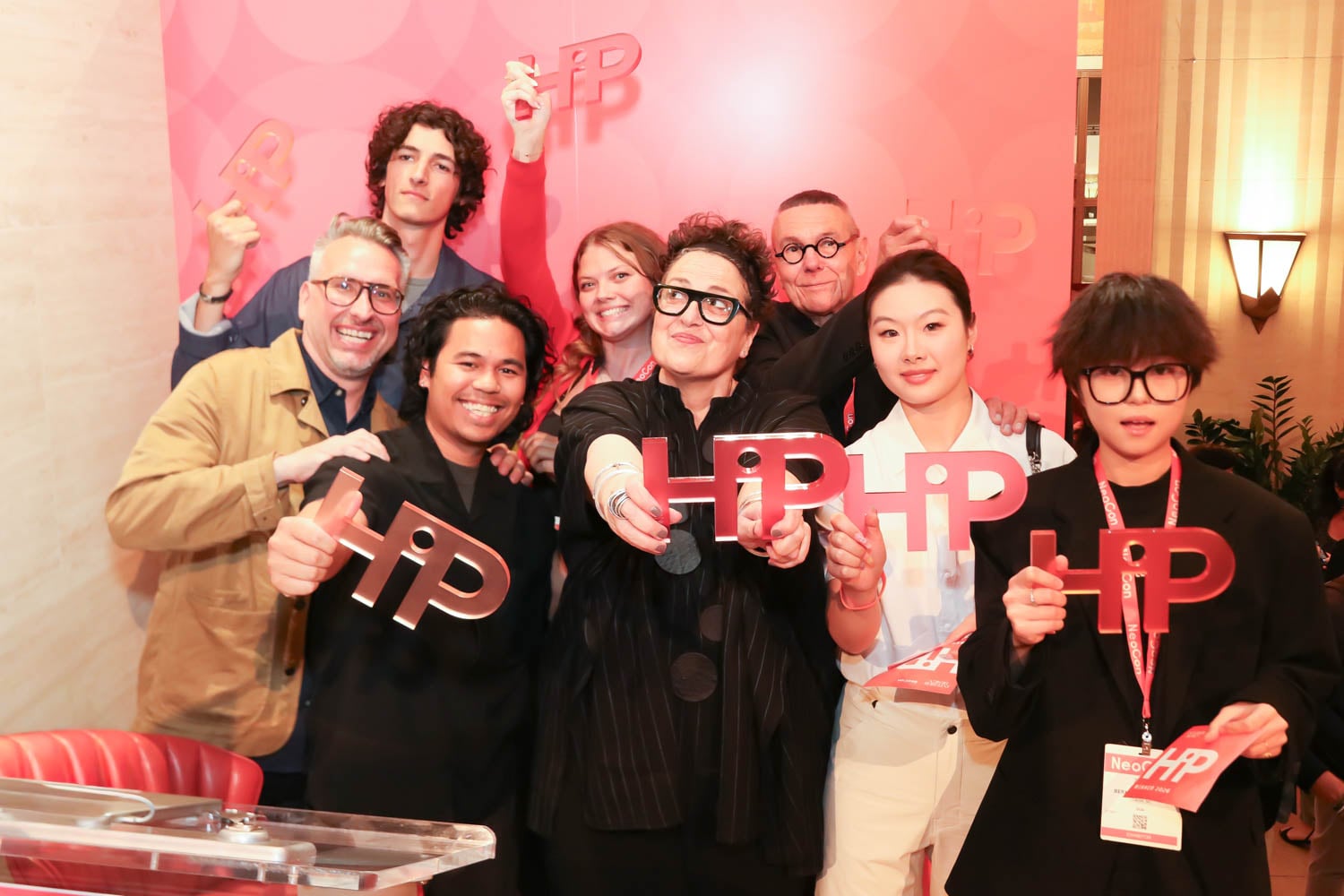 A group of people pose together indoors, smiling and holding large red "HIP" signs at an event in front of a pink and red backdrop.