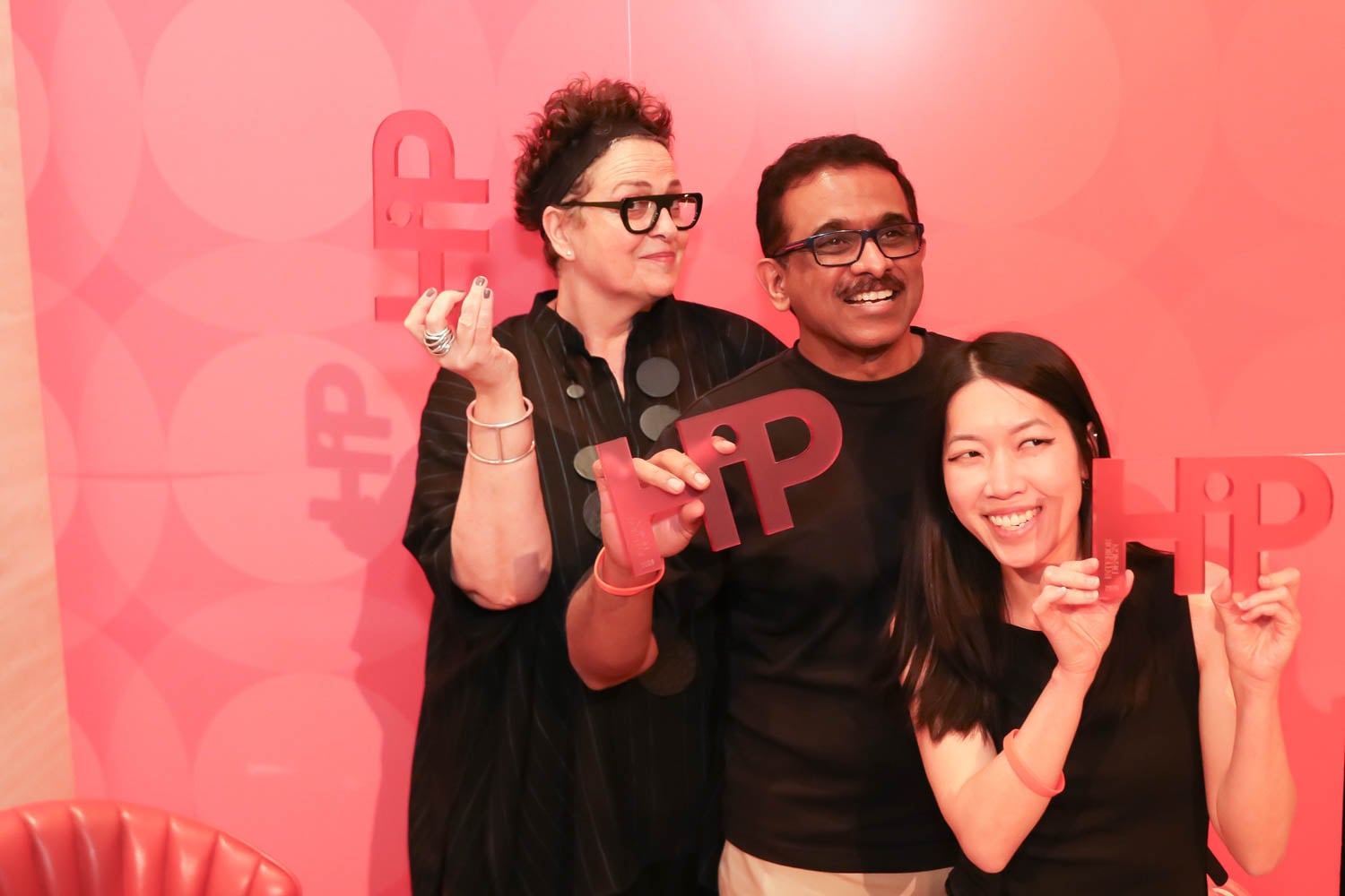 Three people pose in front of a pink backdrop, each holding a red sign with the word "HIP" cut out. They are smiling and appear to be at an event.