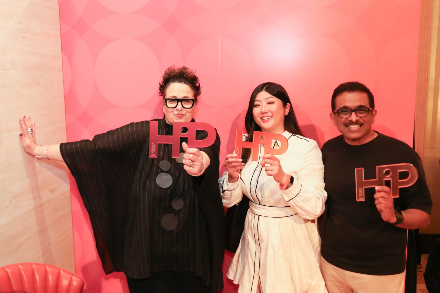 Three people stand in front of a pink backdrop, each holding an award shaped like the letters "HIP" and smiling at the camera.