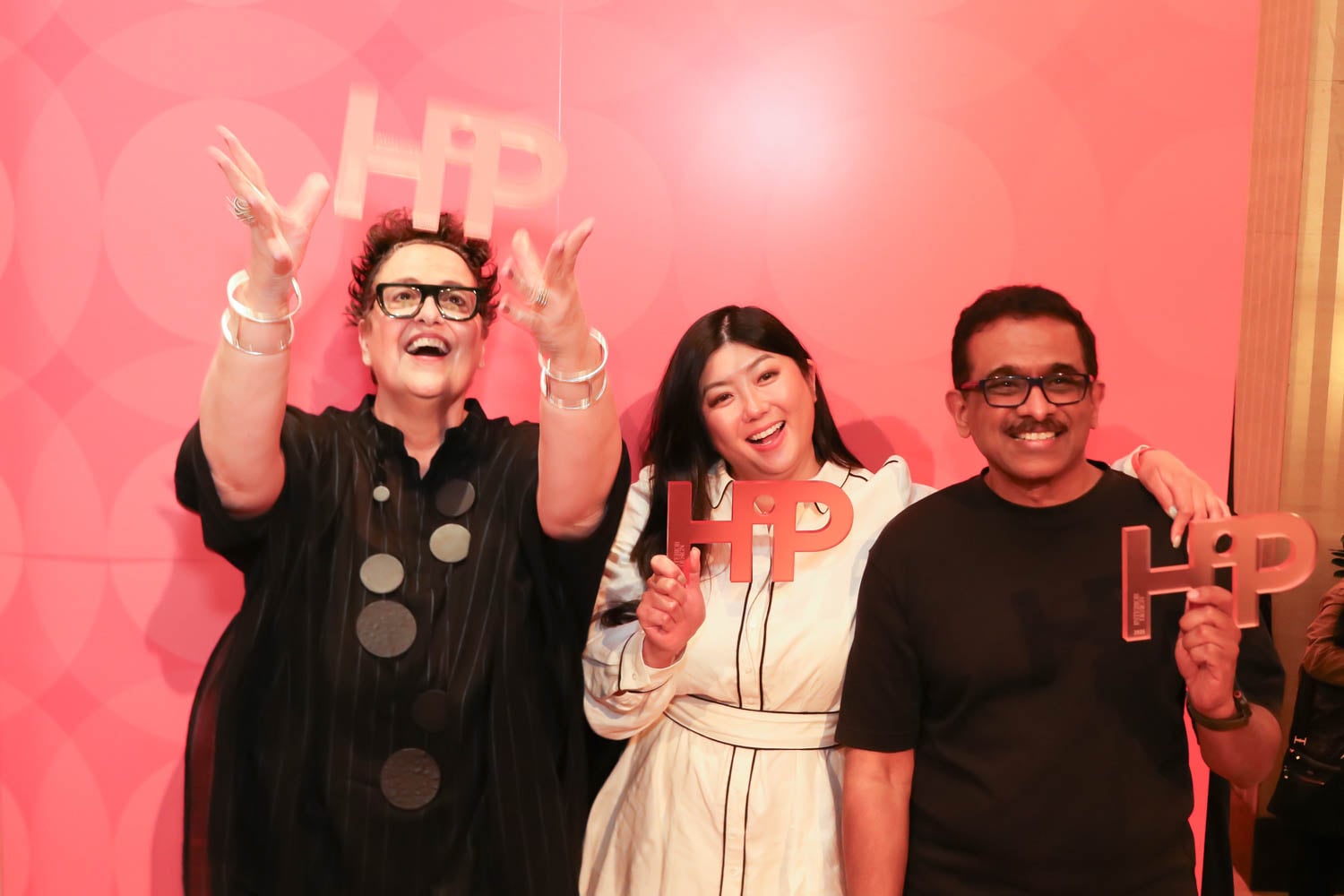 Three people stand in front of a red patterned wall, smiling and holding large "HIP" signs; one person is tossing the sign in the air.