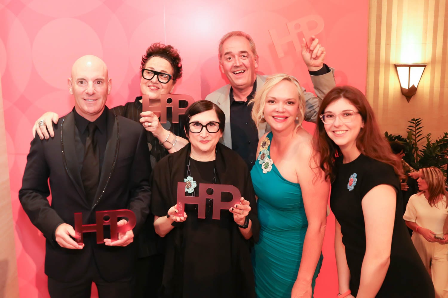 Six people pose together indoors, holding "HiP" awards in front of a pink backdrop with a "HiP" logo. Everyone is smiling and dressed in formal attire.