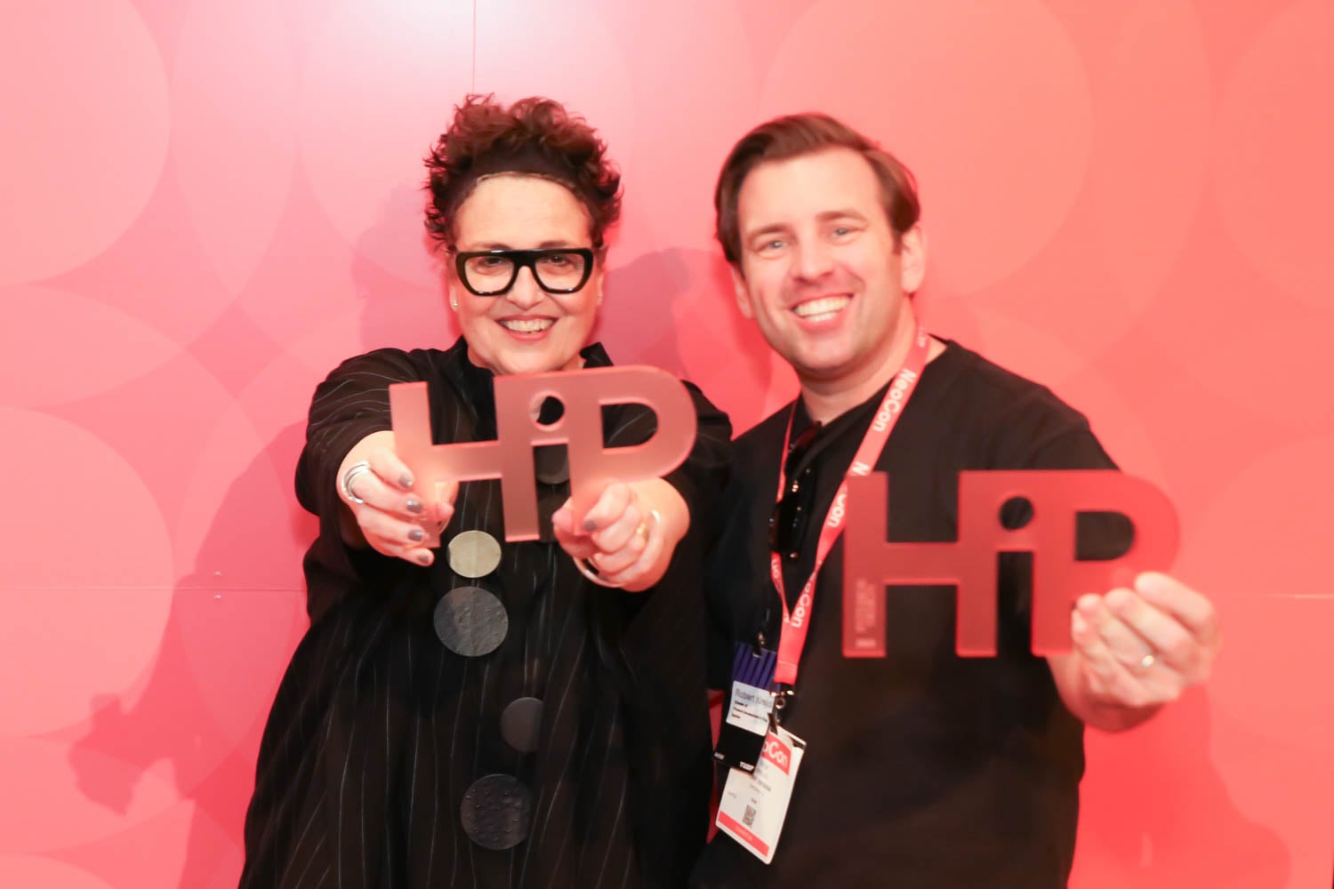 Two people stand in front of a red backdrop, smiling and holding translucent awards shaped with the word "HiP.