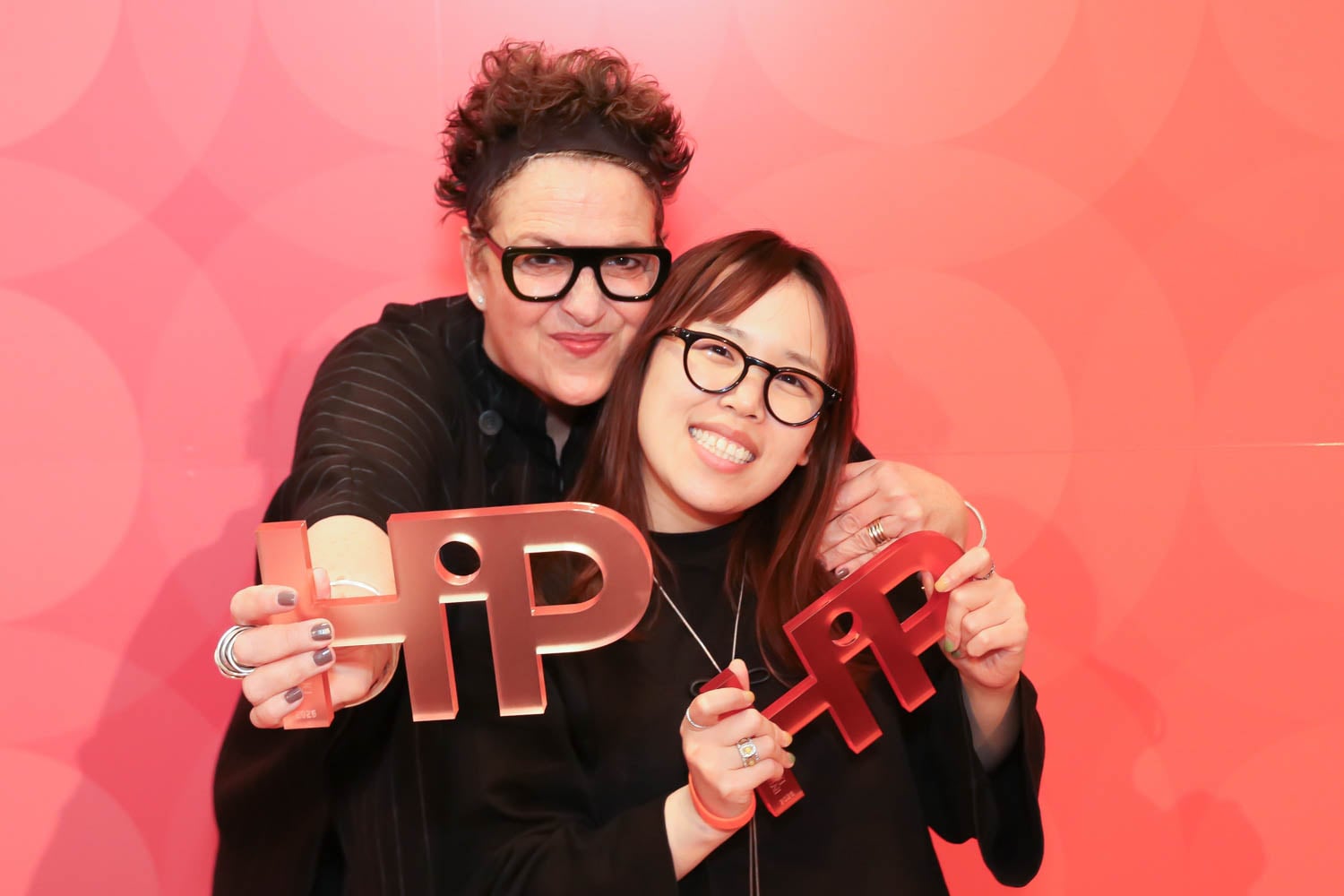 Two people smiling and holding large red "HIP" awards in front of a red background with circular patterns.