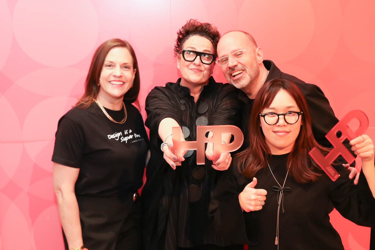 Four people stand in front of a pink backdrop, two holding large "HP" letter props and one giving a thumbs up, all smiling at the camera.