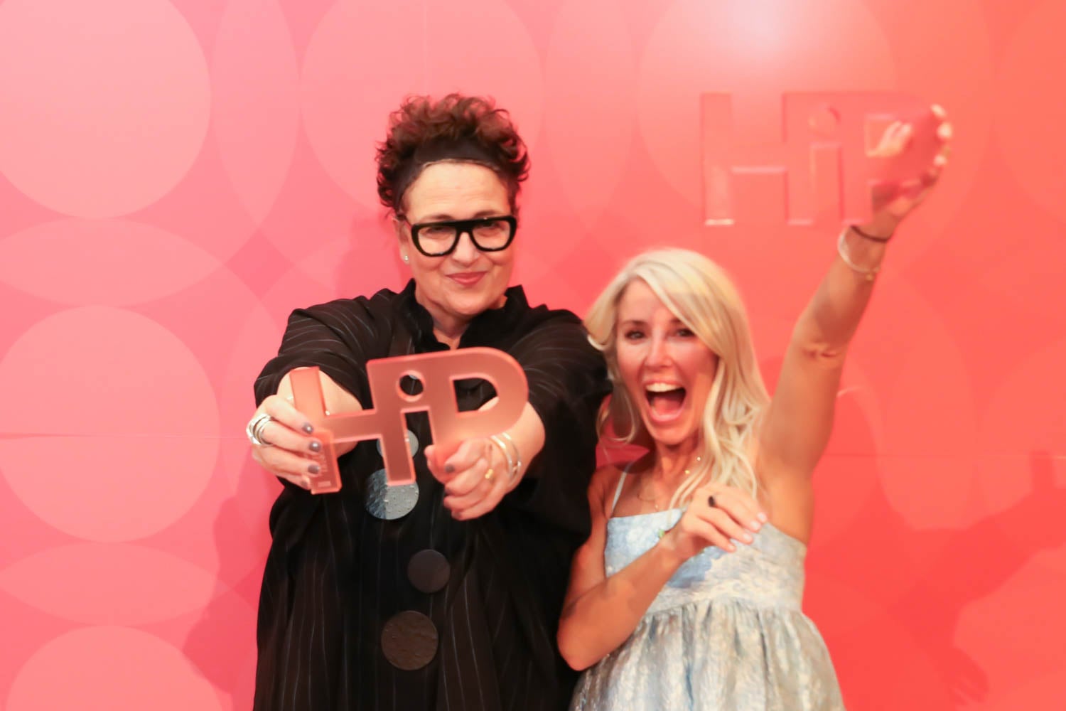 Two women stand in front of a pink backdrop, smiling and holding up “HIP” shaped awards toward the camera.