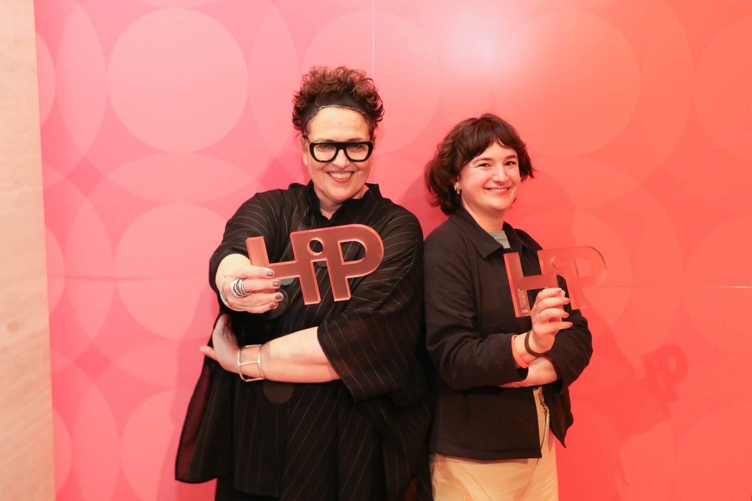 Two people stand in front of a pink patterned wall, each holding a “HiP” award and smiling at the camera.