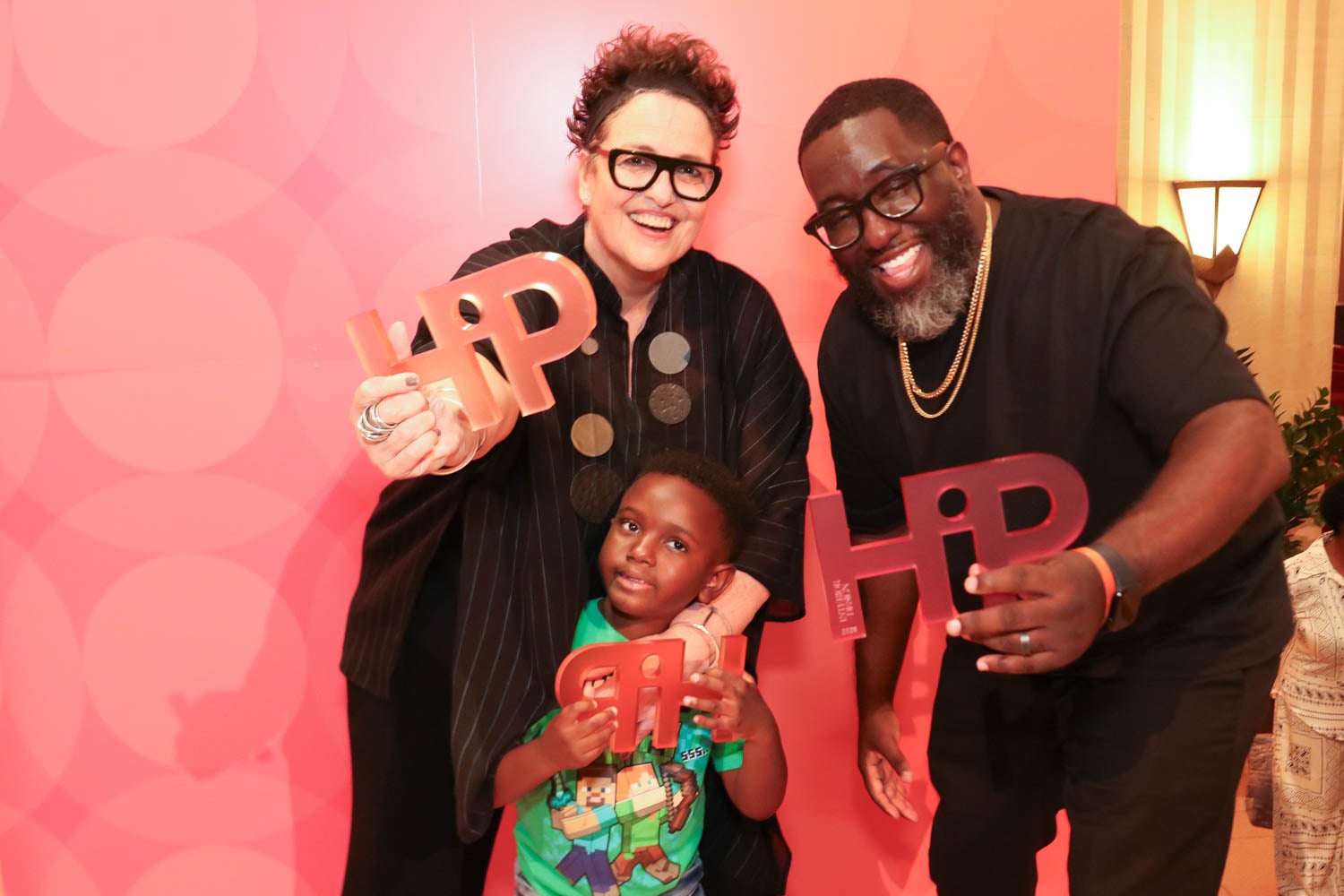 Three people, two adults and one child, smile and hold large, stylized “Hip” signs in front of a pink backdrop with circular patterns.