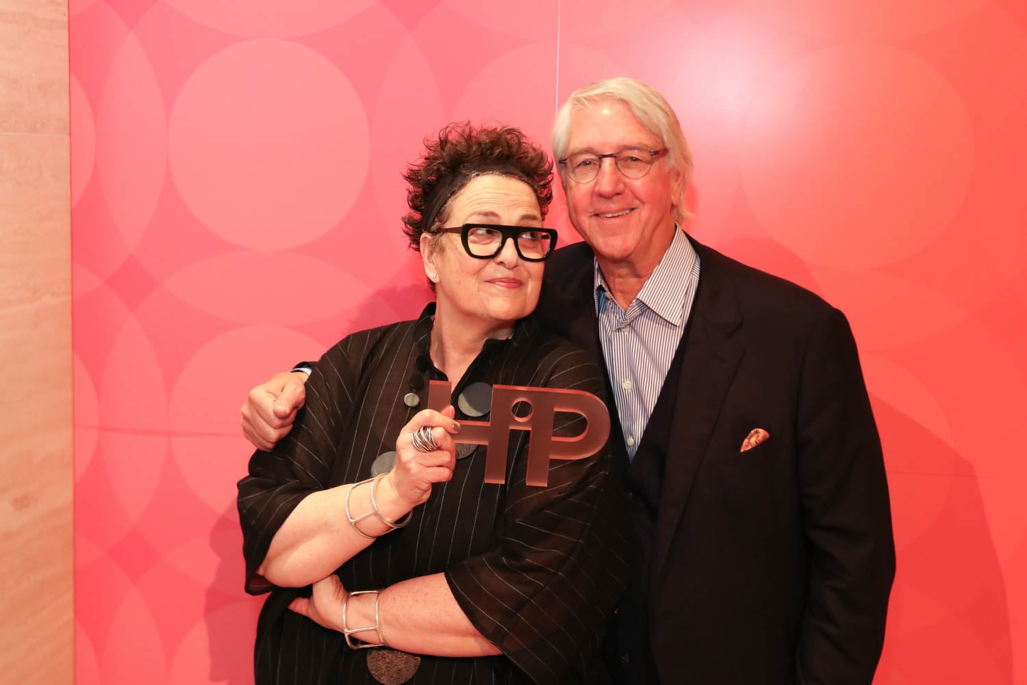 Two people stand together in front of a red patterned wall; one holds a large metallic key-shaped object and both are smiling.