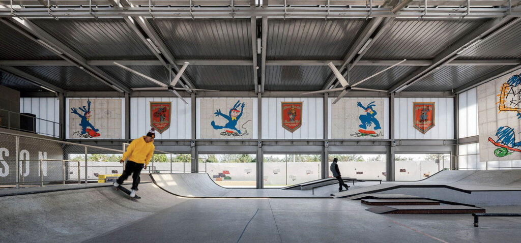 Two people skateboarding in an indoor skate park with metal beams and colorful wall art depicting cartoon characters and shields.