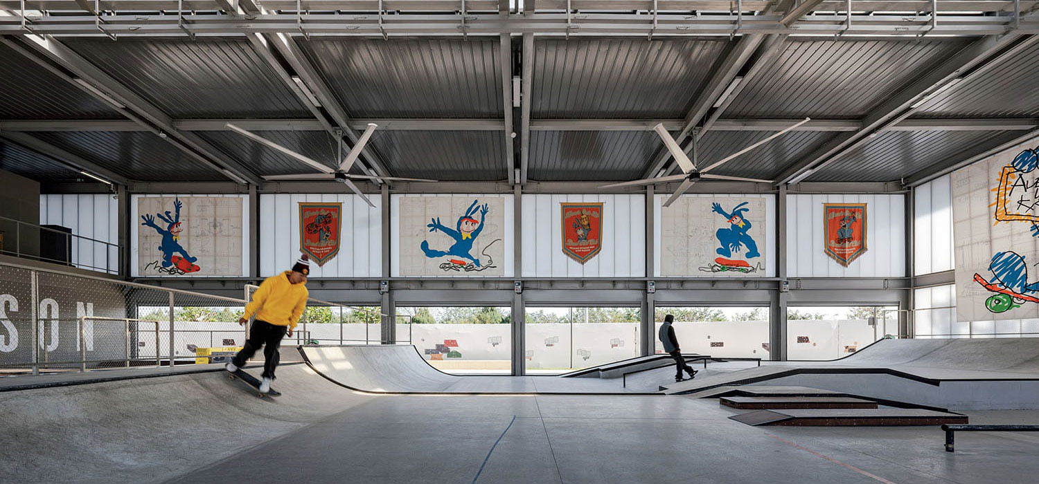 Two people skateboarding in an indoor skate park with metal beams and colorful wall art depicting cartoon characters and shields.