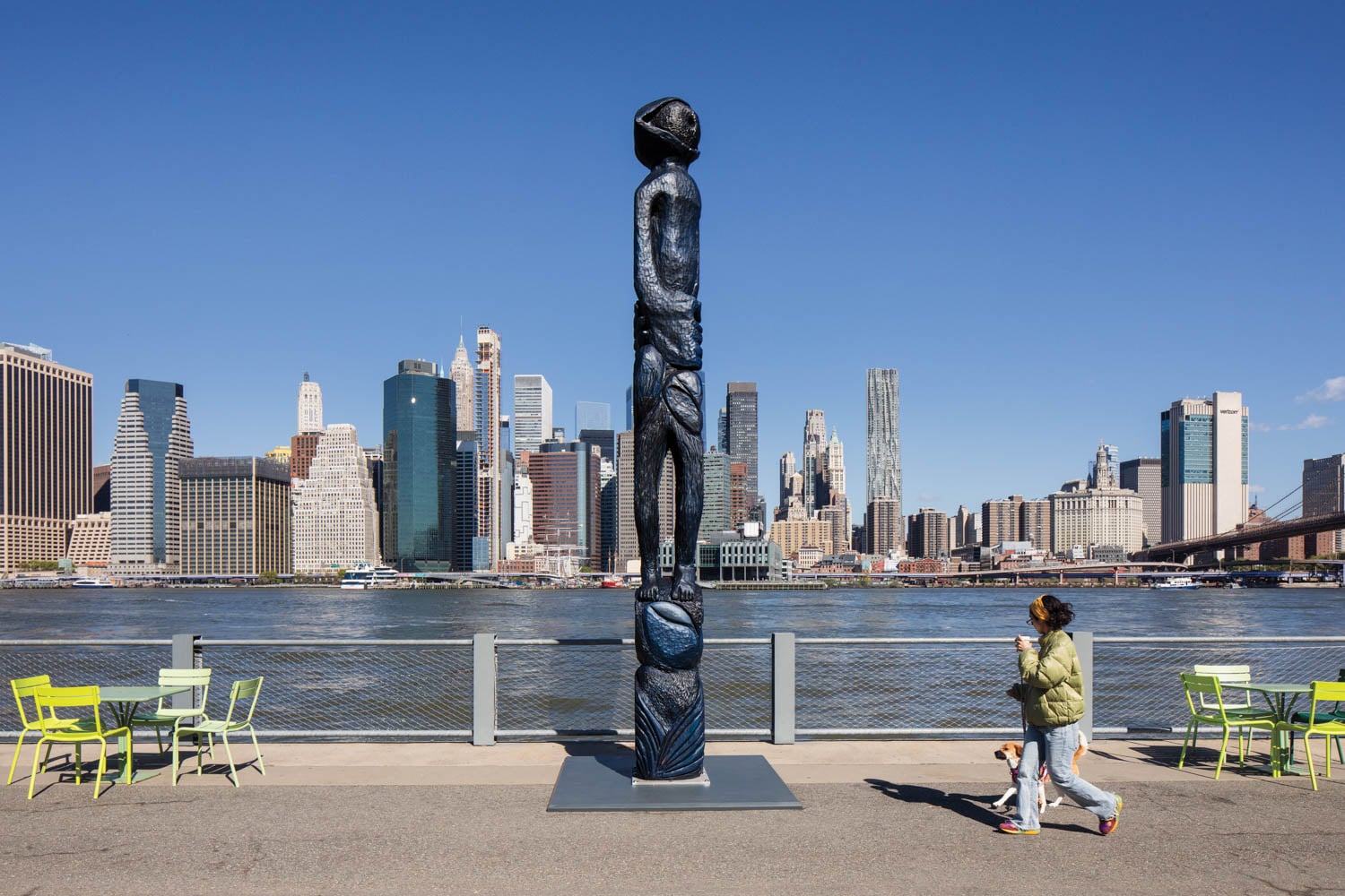 A tall, dark statue of a standing figure overlooks the East River with the Manhattan skyline in the background; a person walks by with a drink and dog.