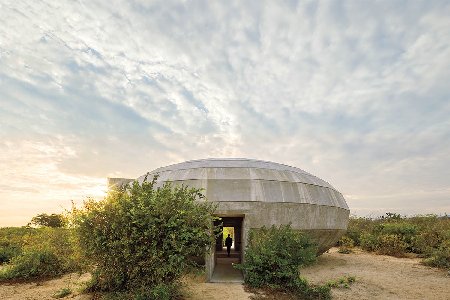 Round, bunker-like concrete building with an oval shape set in a natural landscape under a partly cloudy sky, with a person visible through the entrance.