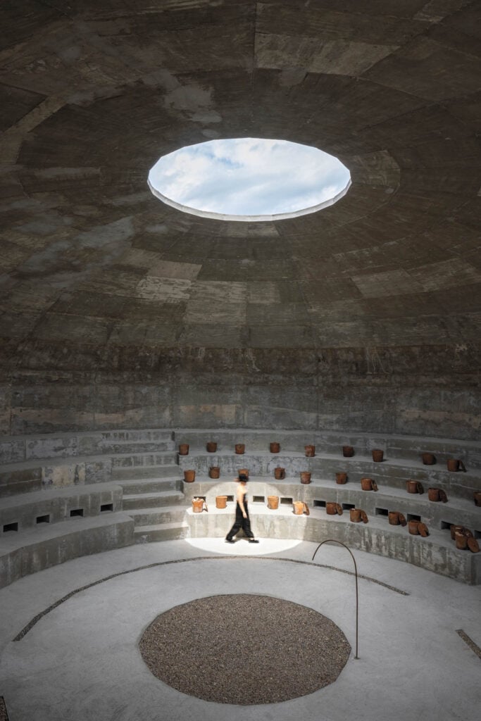 Person walking inside a circular, concrete amphitheater with tiered seating and a large round skylight in the ceiling, allowing natural light to enter.