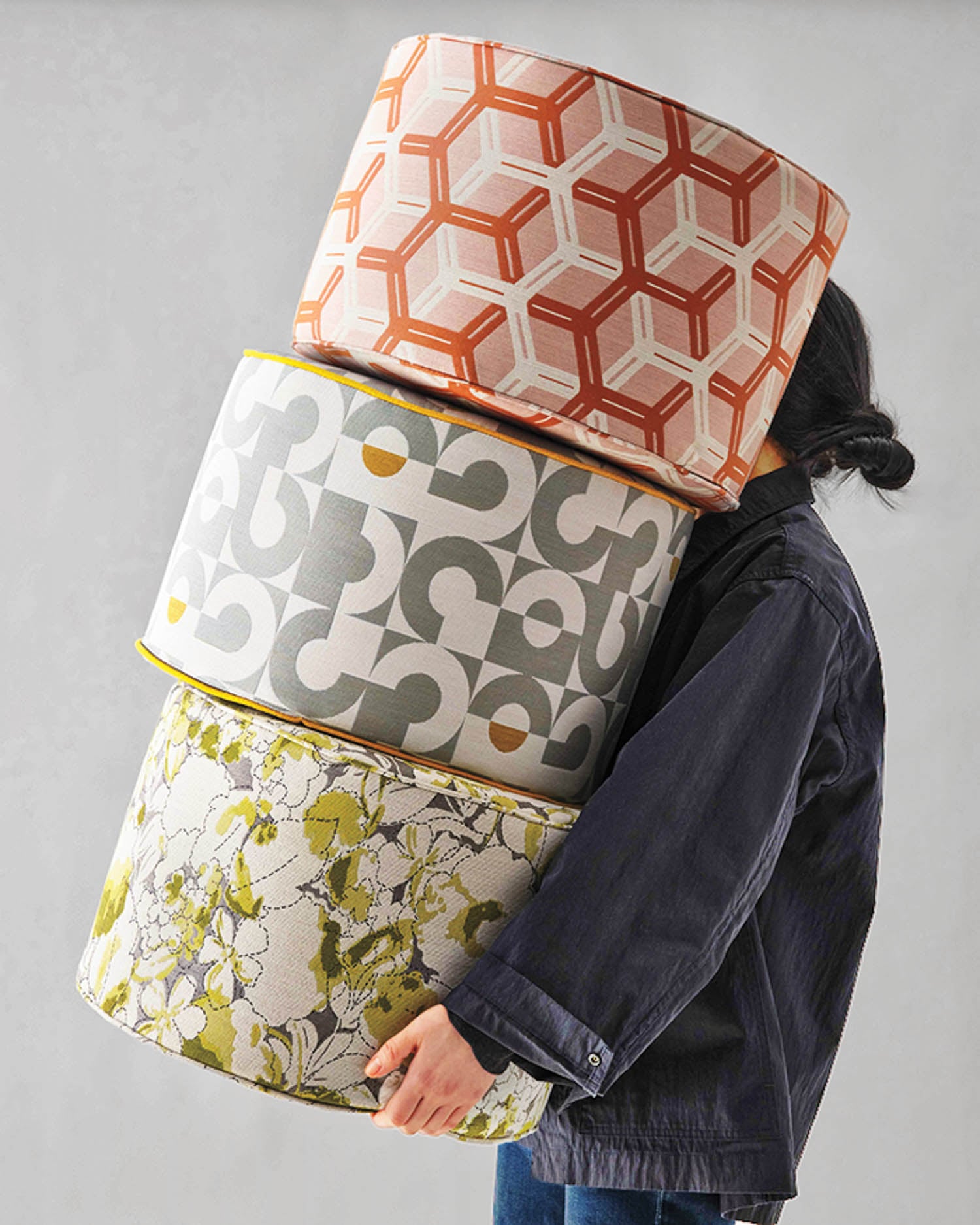 A person carries three large, patterned cylindrical cushions stacked on top of each other against a plain background.