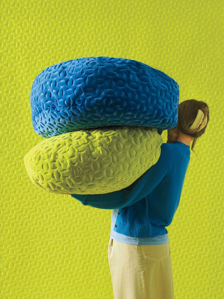 A person in a blue sweater and beige pants holds two large, textured cushions—one blue and one yellow—against a bright green textured background.