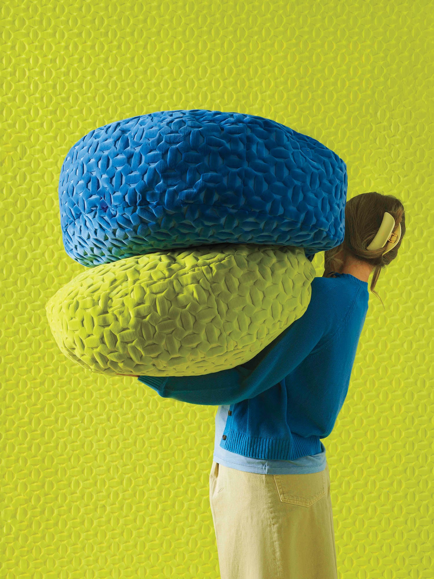 A person in a blue sweater and beige pants holds two large, textured cushions—one blue and one yellow—against a bright green textured background.