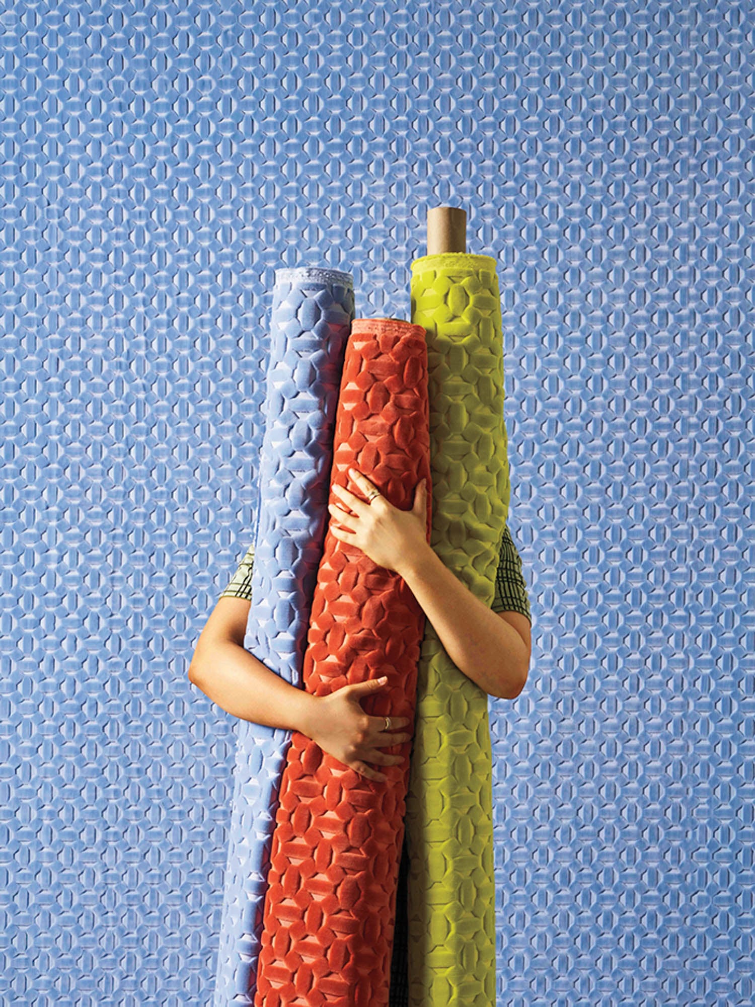 A person stands against a blue patterned wall, holding three large rolls of fabric in blue, red, and yellow, partially covering their body and face.