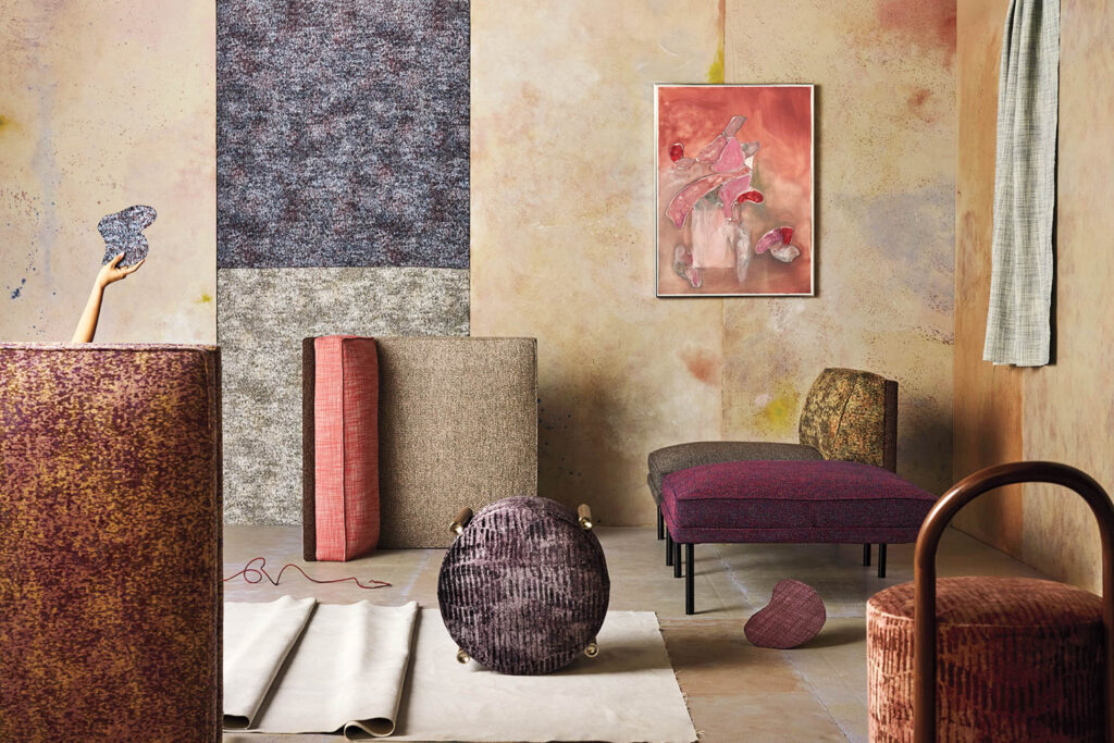 A modern room with textured furniture in muted tones, abstract wall art, a hand holding a textured object, and a partially rolled-up rug on the floor.
