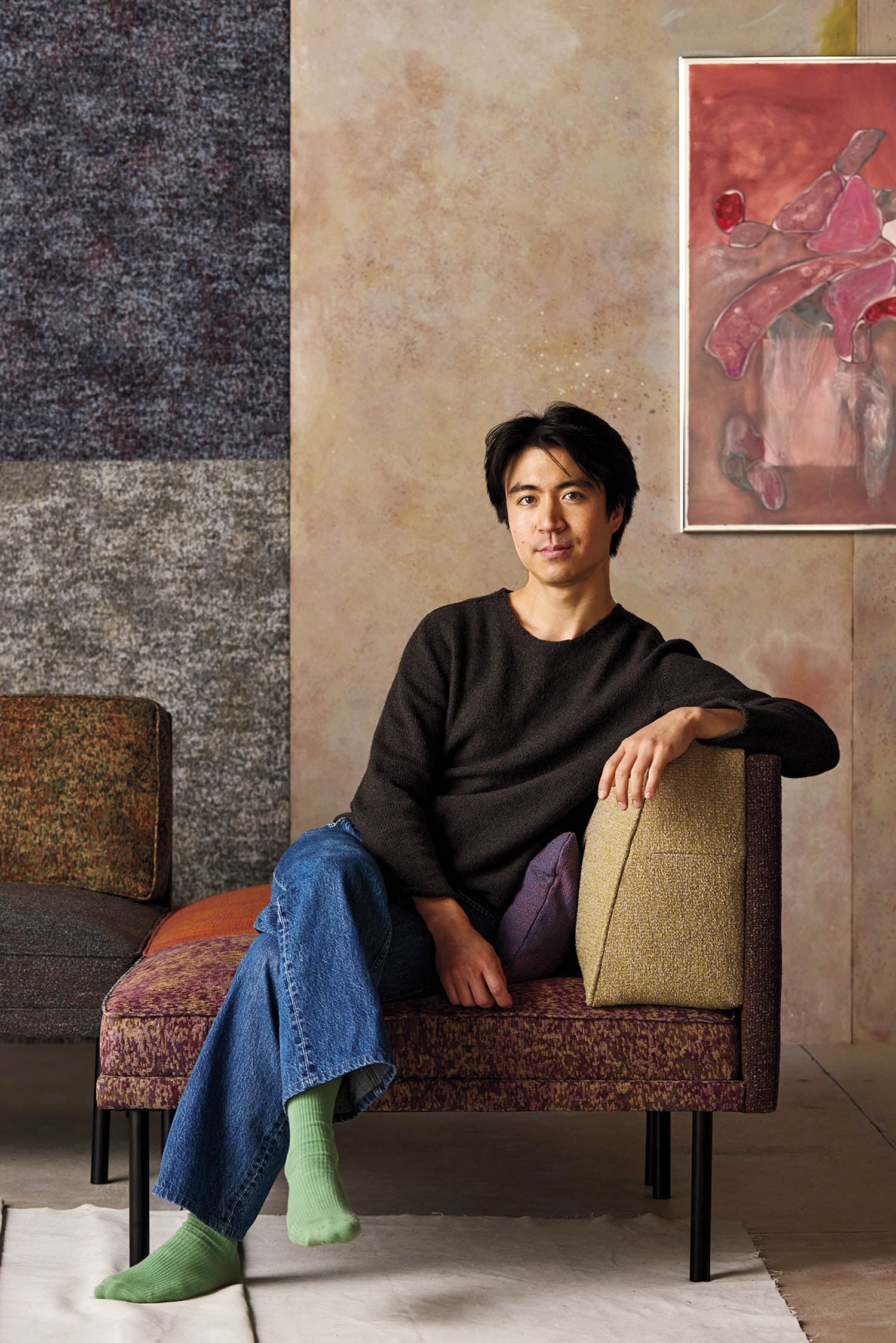 A person with dark hair, wearing a black sweater, blue jeans, and green socks, sits on a modern, patterned chair in a room with textured walls and abstract artwork.