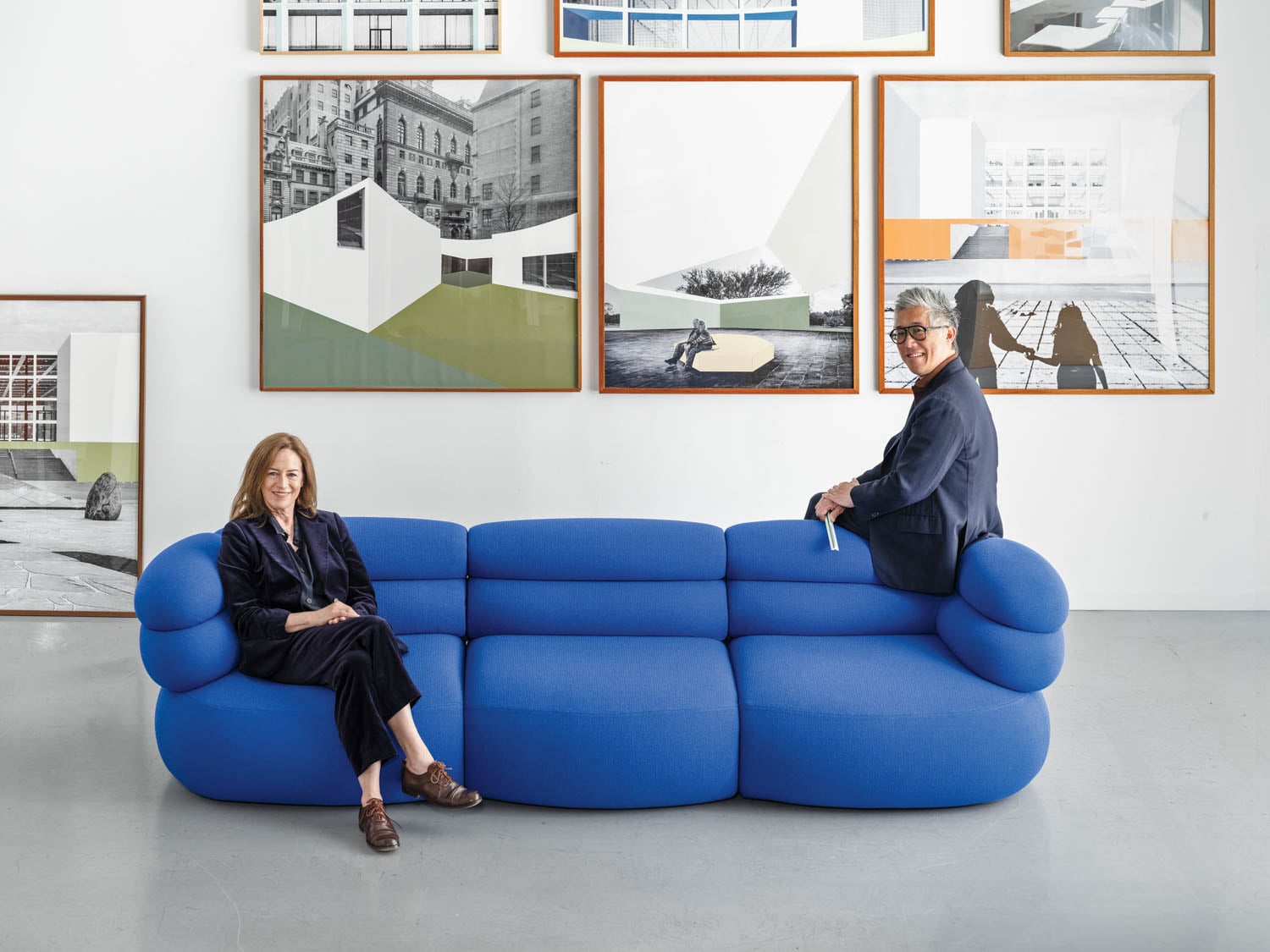 Two people sit at opposite ends of a modern blue sofa in a gallery space with architectural artworks displayed on the wall behind them.