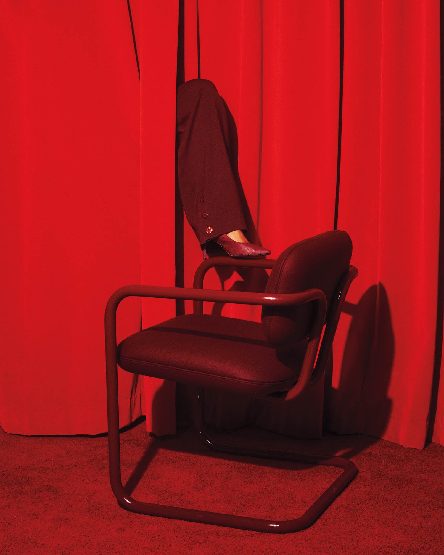 A single leg wearing dark dress pants and a black shoe extends through red curtains above a red chair on a red carpeted floor.